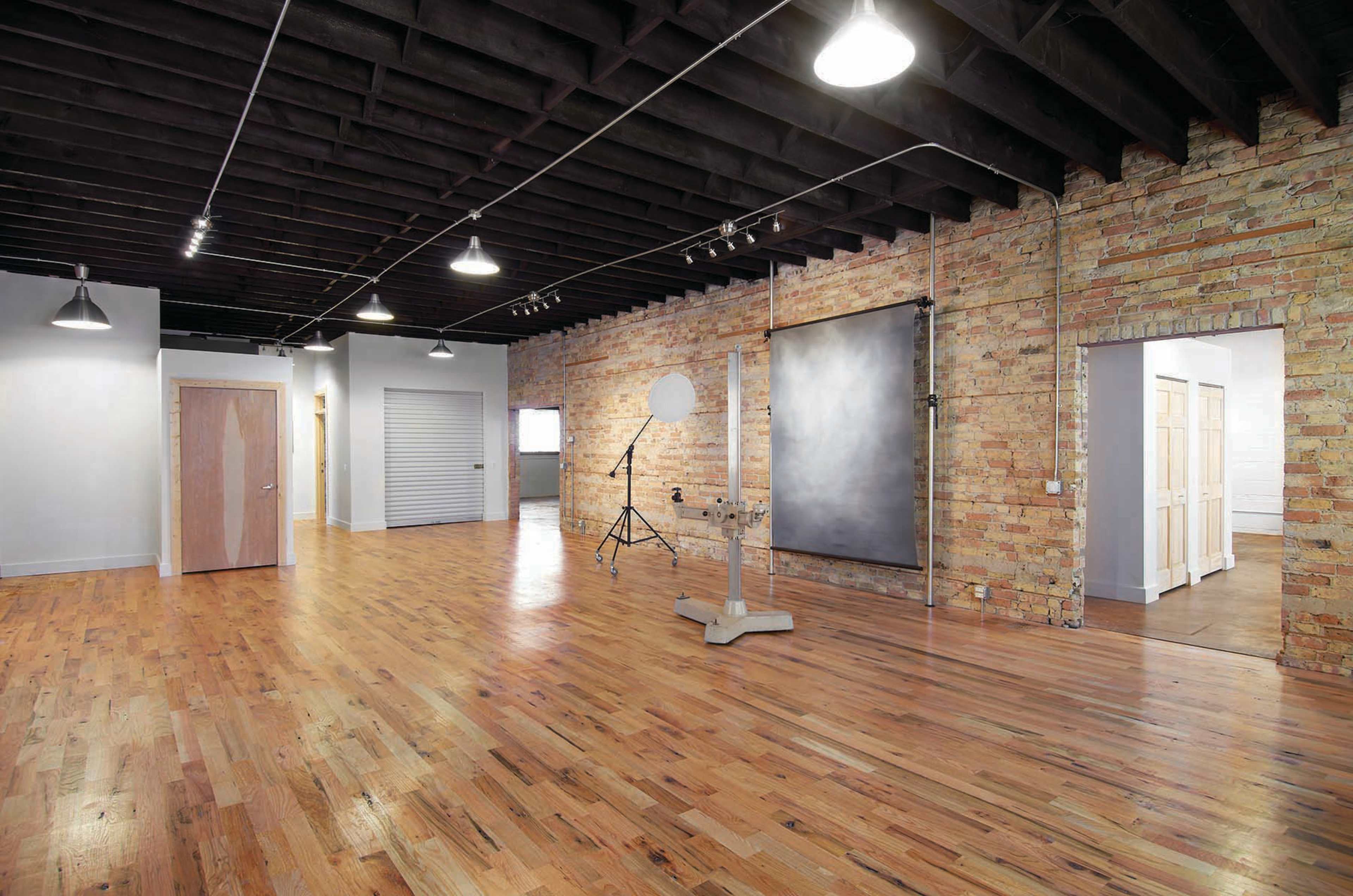 A spacious interior room with hardwood flooring, exposed brick walls, and overhead lighting fixtures.