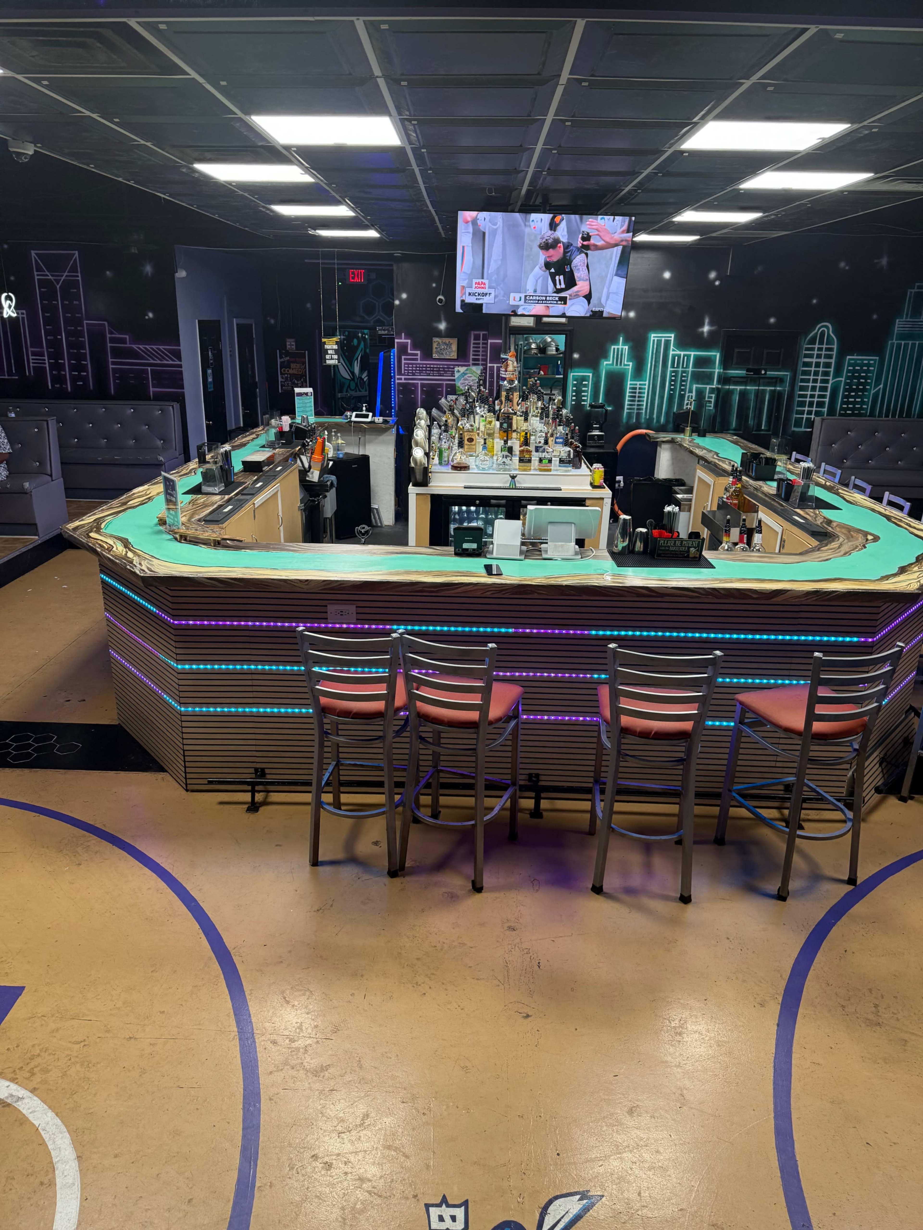 The image shows a modern bar area with colorful LED lighting and a large television displaying a sports game, surrounded by high stools.