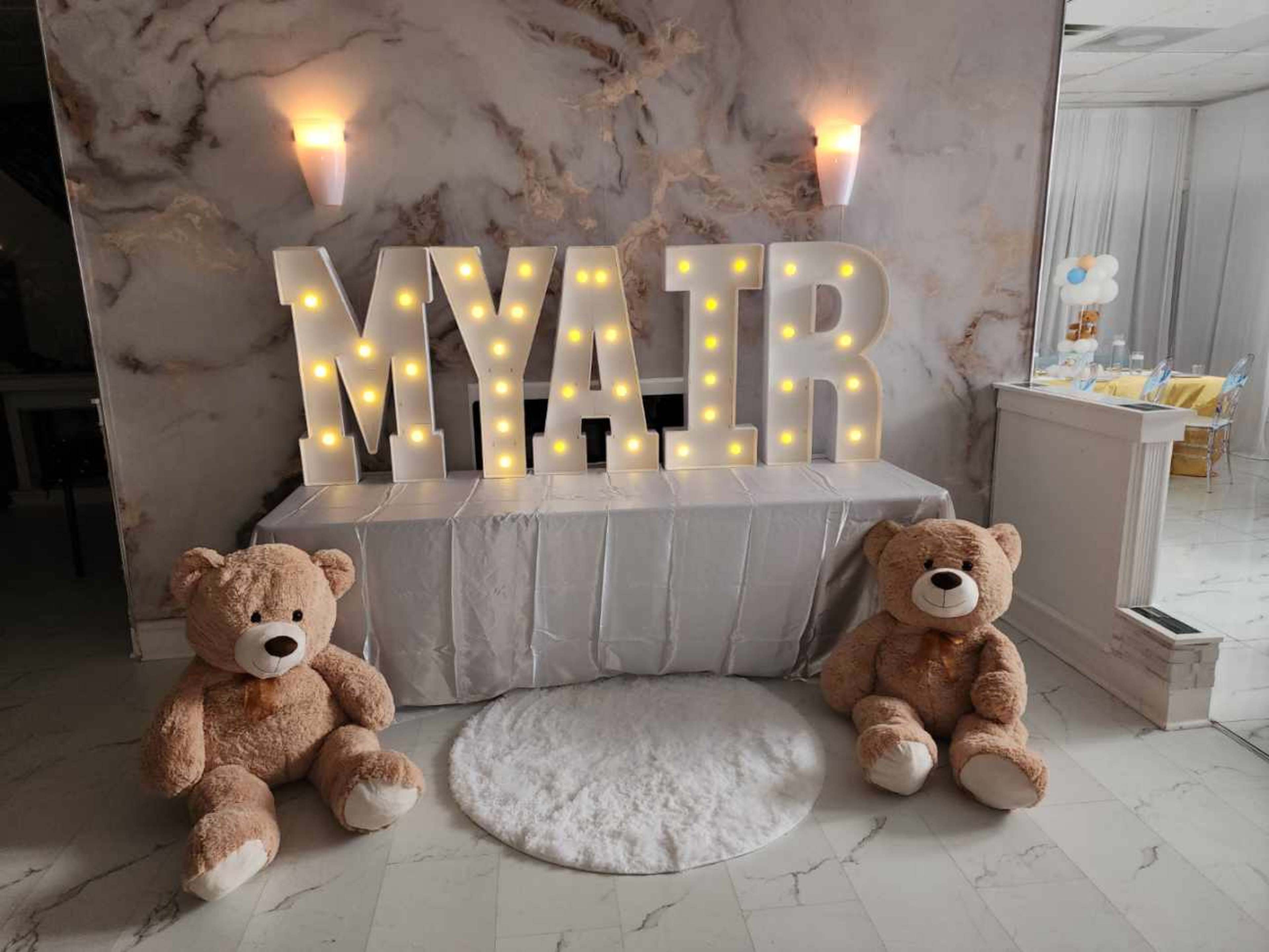 The scene features large, illuminated letters spelling "MYAIR" displayed on a table, accompanied by two large teddy bears sitting on either side on a white rug.