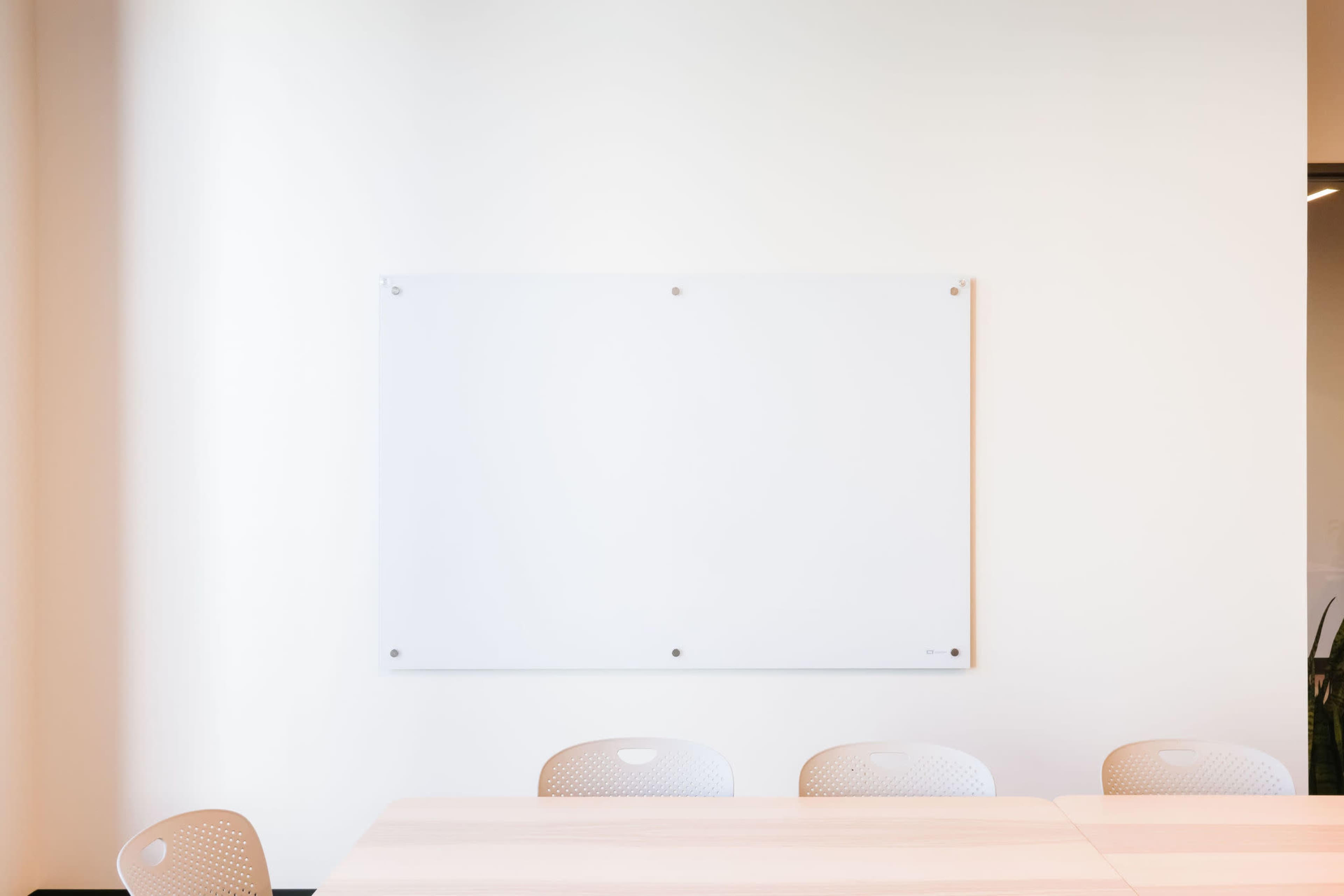 A blank whiteboard is mounted on a light-colored wall above a wooden conference table with several empty chairs around it.