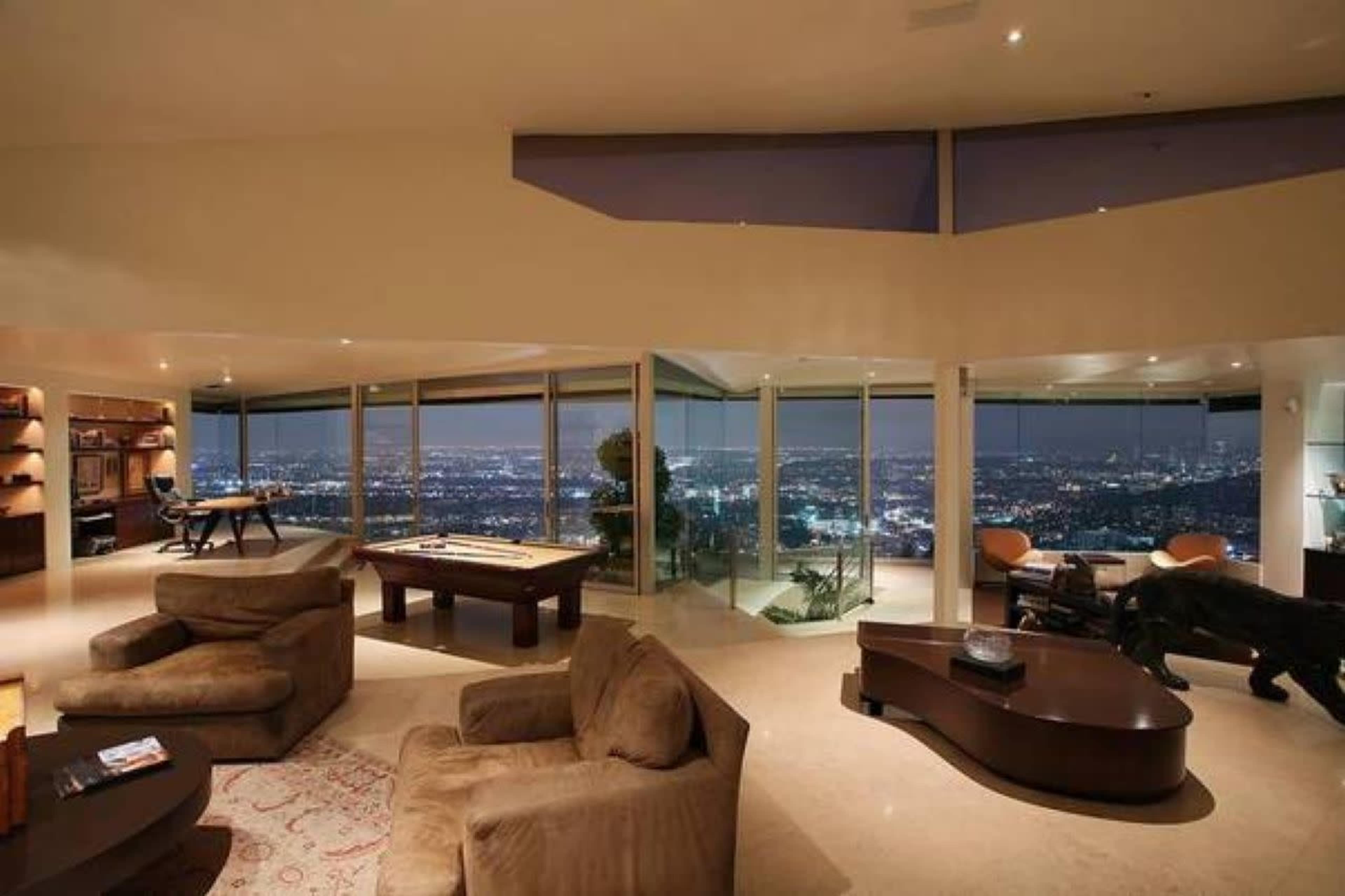The image depicts a modern, spacious living room with large windows offering a panoramic view of a brightly lit city at night.