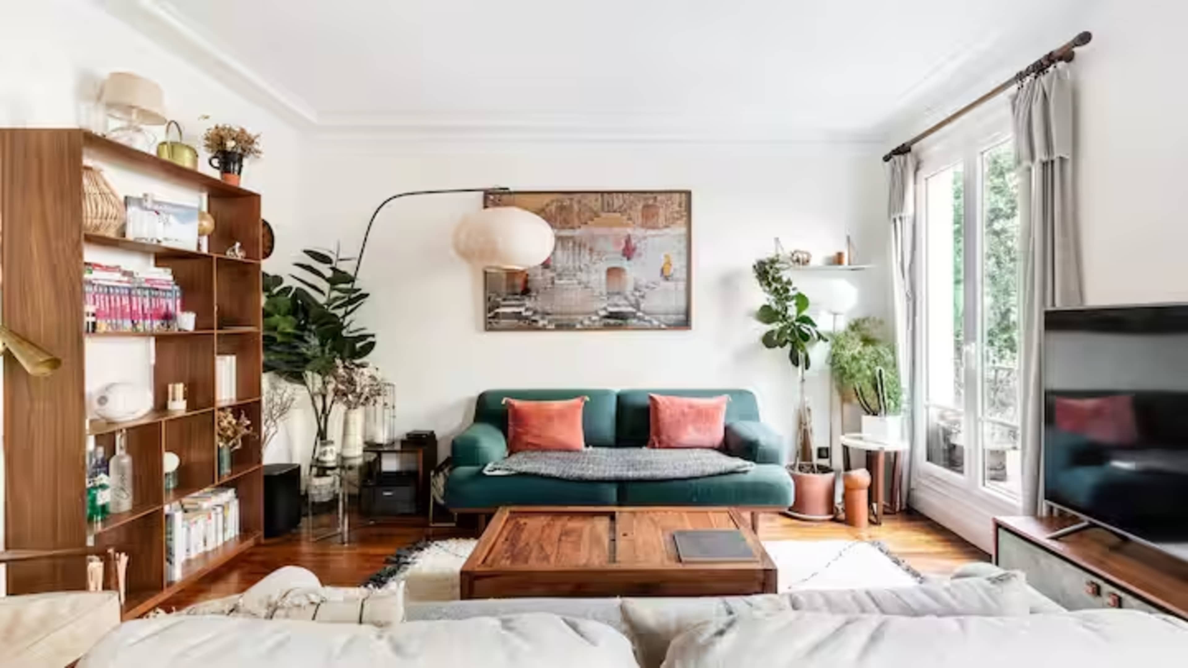 The image shows a bright living room featuring a green sofa, a wooden coffee table, a large bookshelf, and a floor lamp, with large windows allowing natural light to enter.
