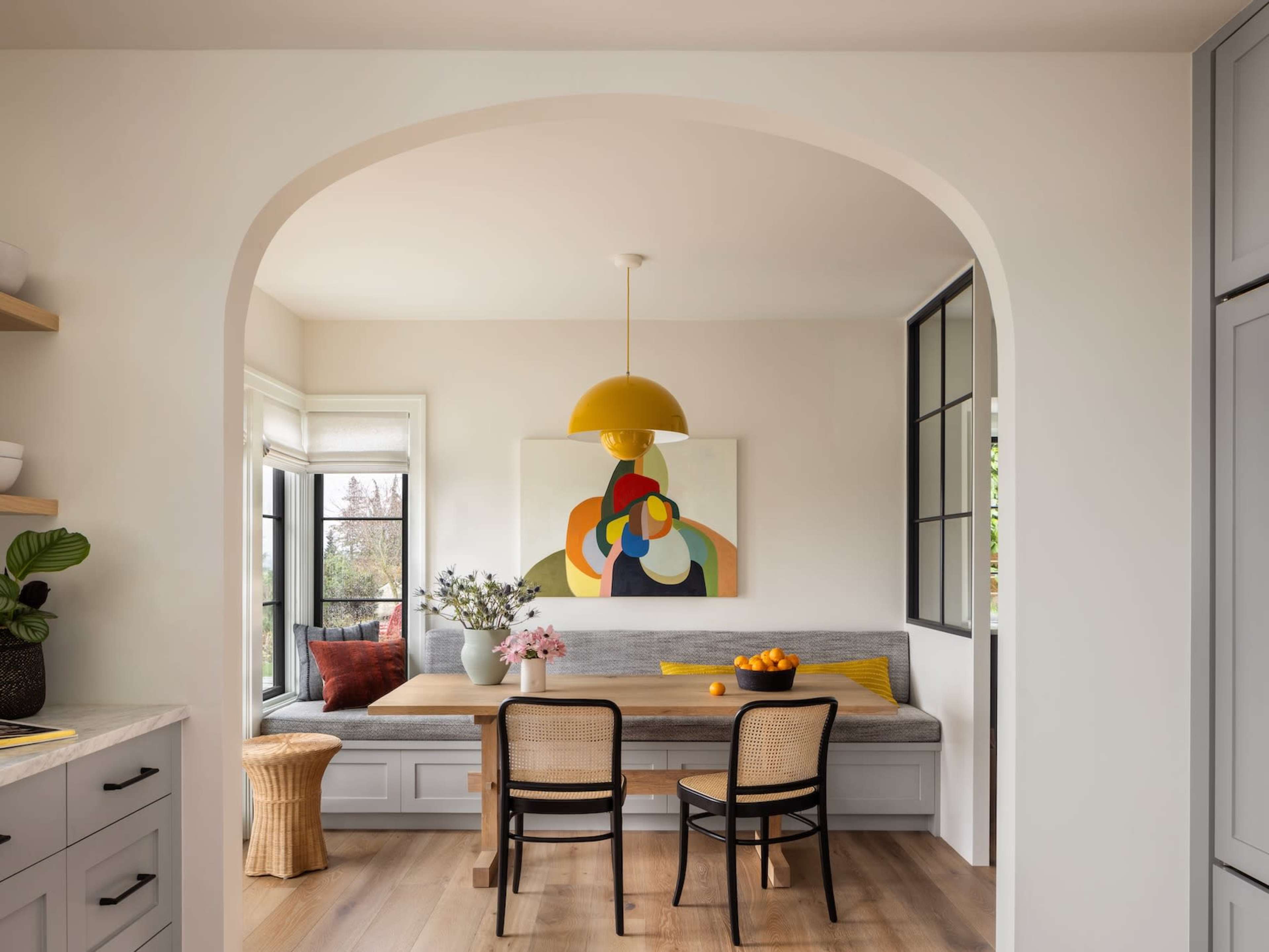 A bright dining nook features a round table surrounded by chairs, with a colorful artwork hanging on the wall and a cozy bench seat along the window.
