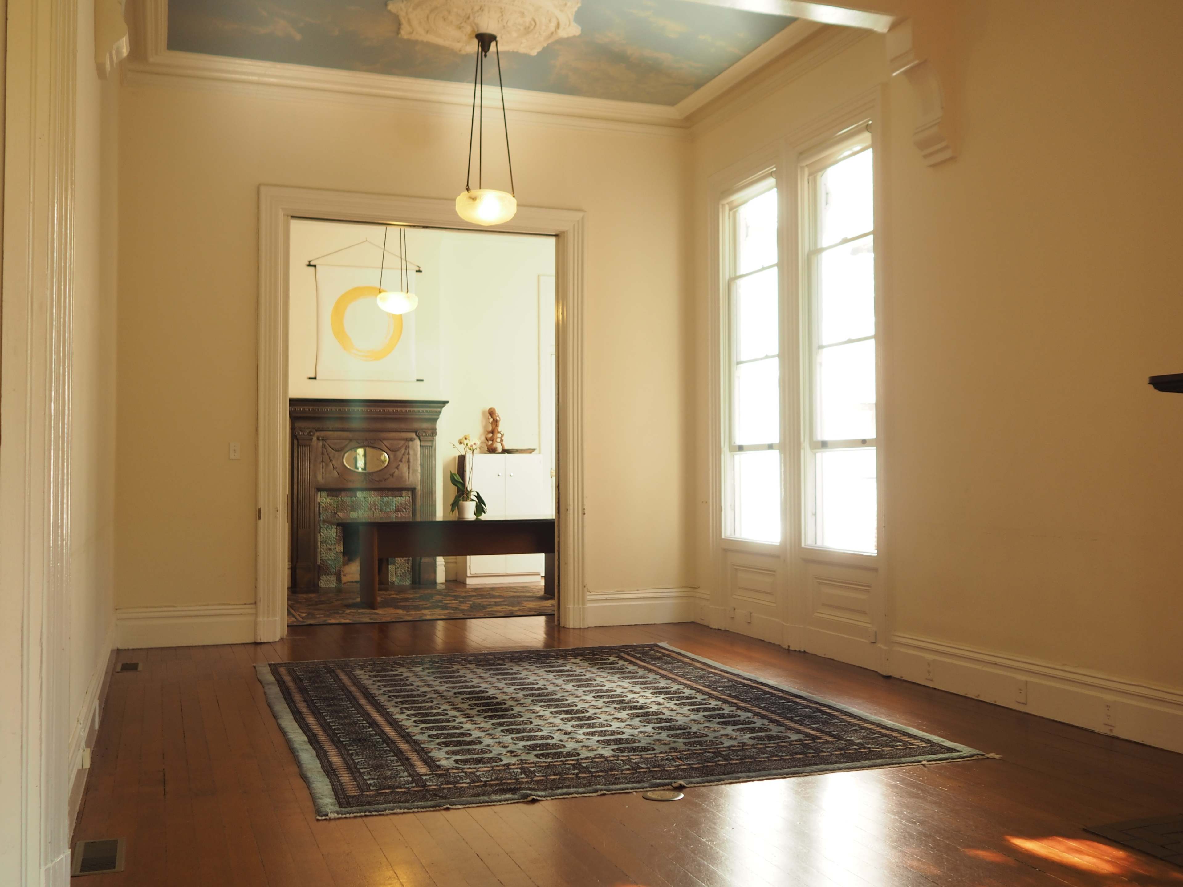 A spacious interior room with a wooden floor, an area rug, and large windows, leading to another room with a decorative fireplace.