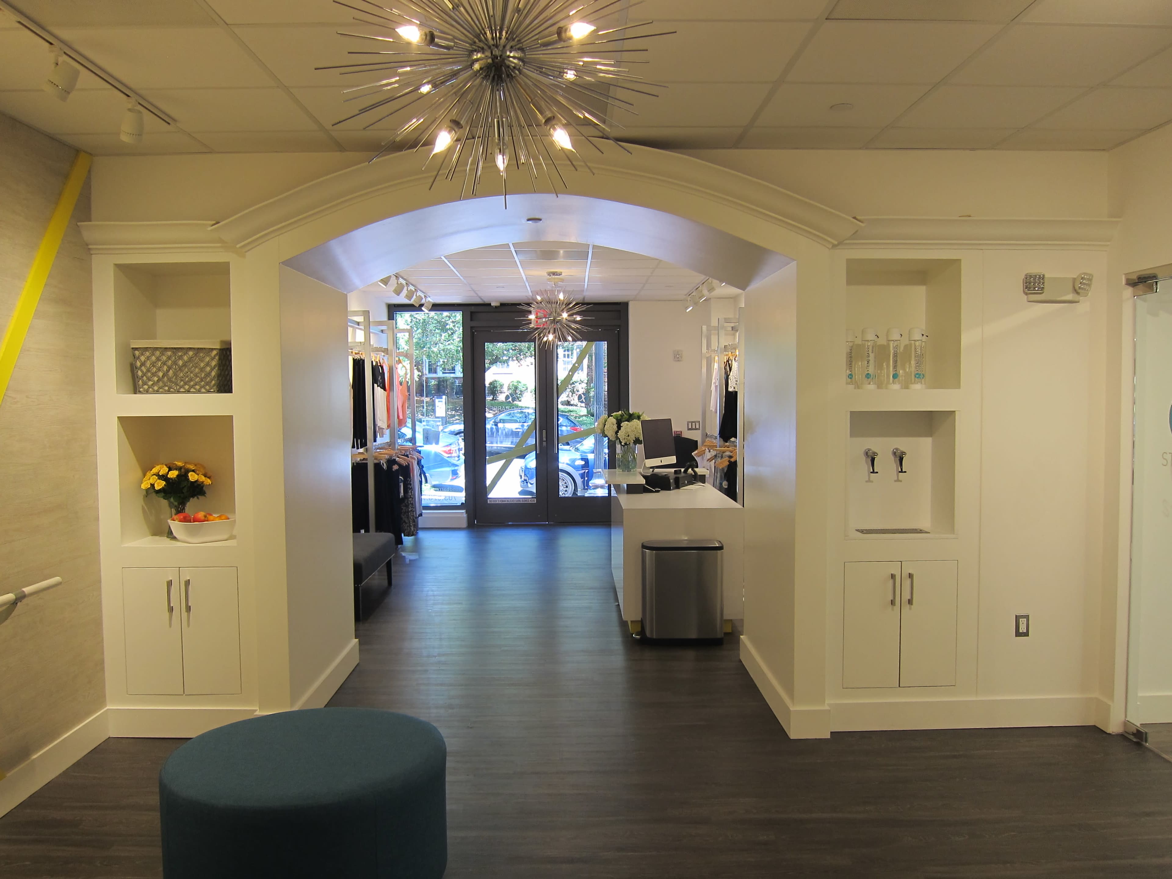 The image shows a modern reception area with a hallway leading to a front entrance, featuring light fixtures and display shelves on either side.