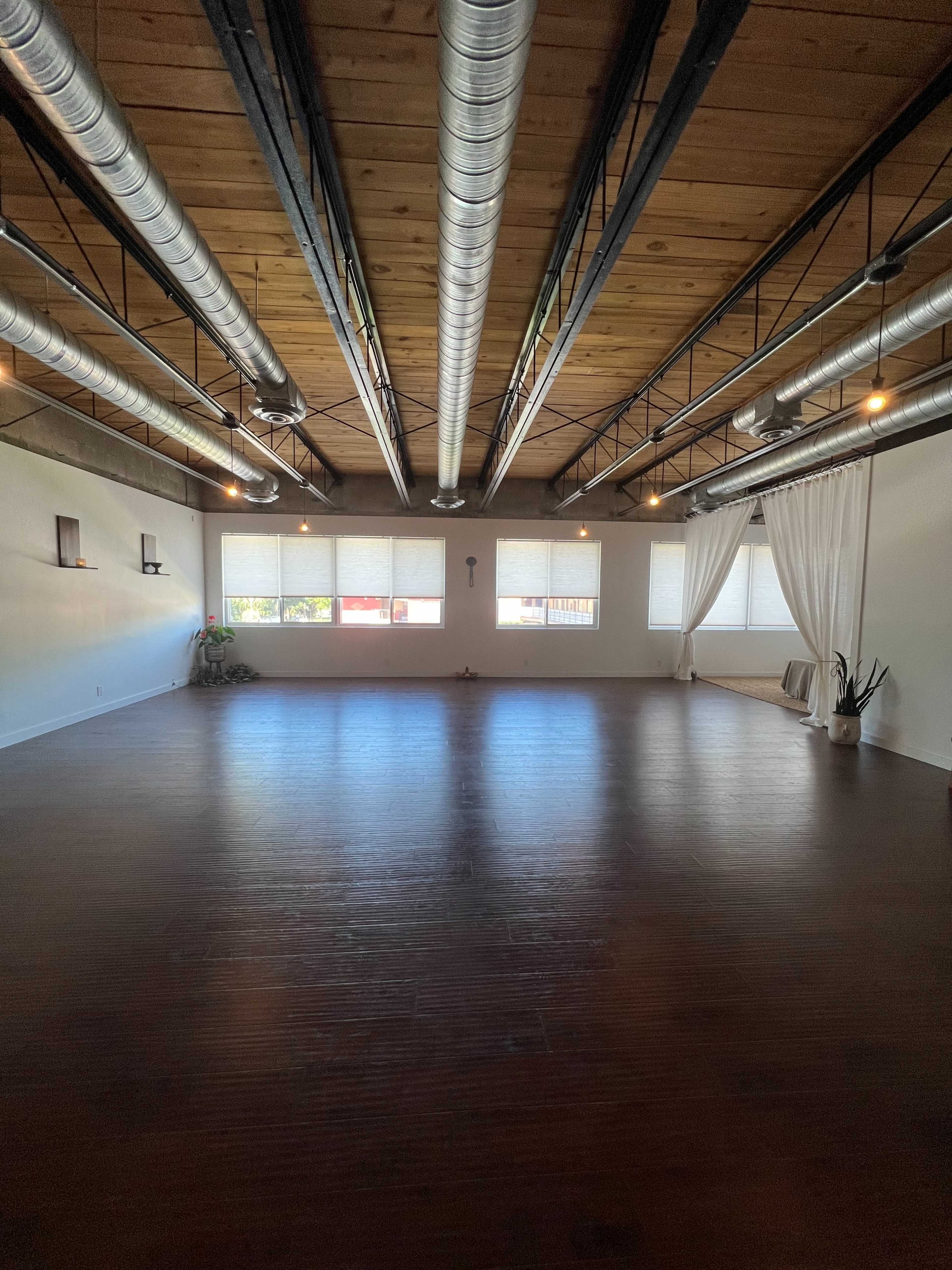 An empty indoor space with a wooden floor, large windows, and exposed metal ductwork on the ceiling.