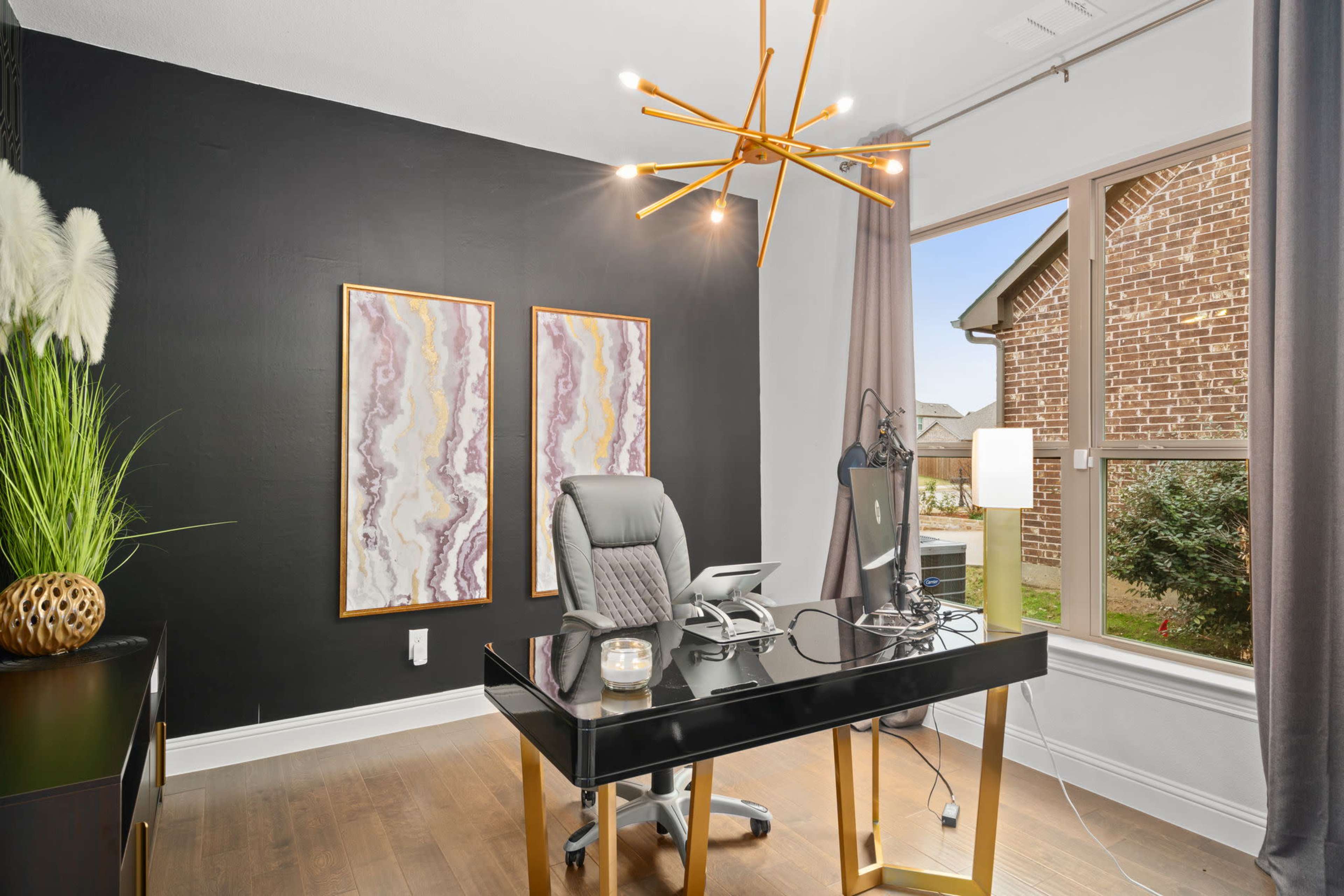 A modern office features a black accent wall, two colorful framed artworks, a stylish desk with a chair, and a large window overlooking an outdoor view.