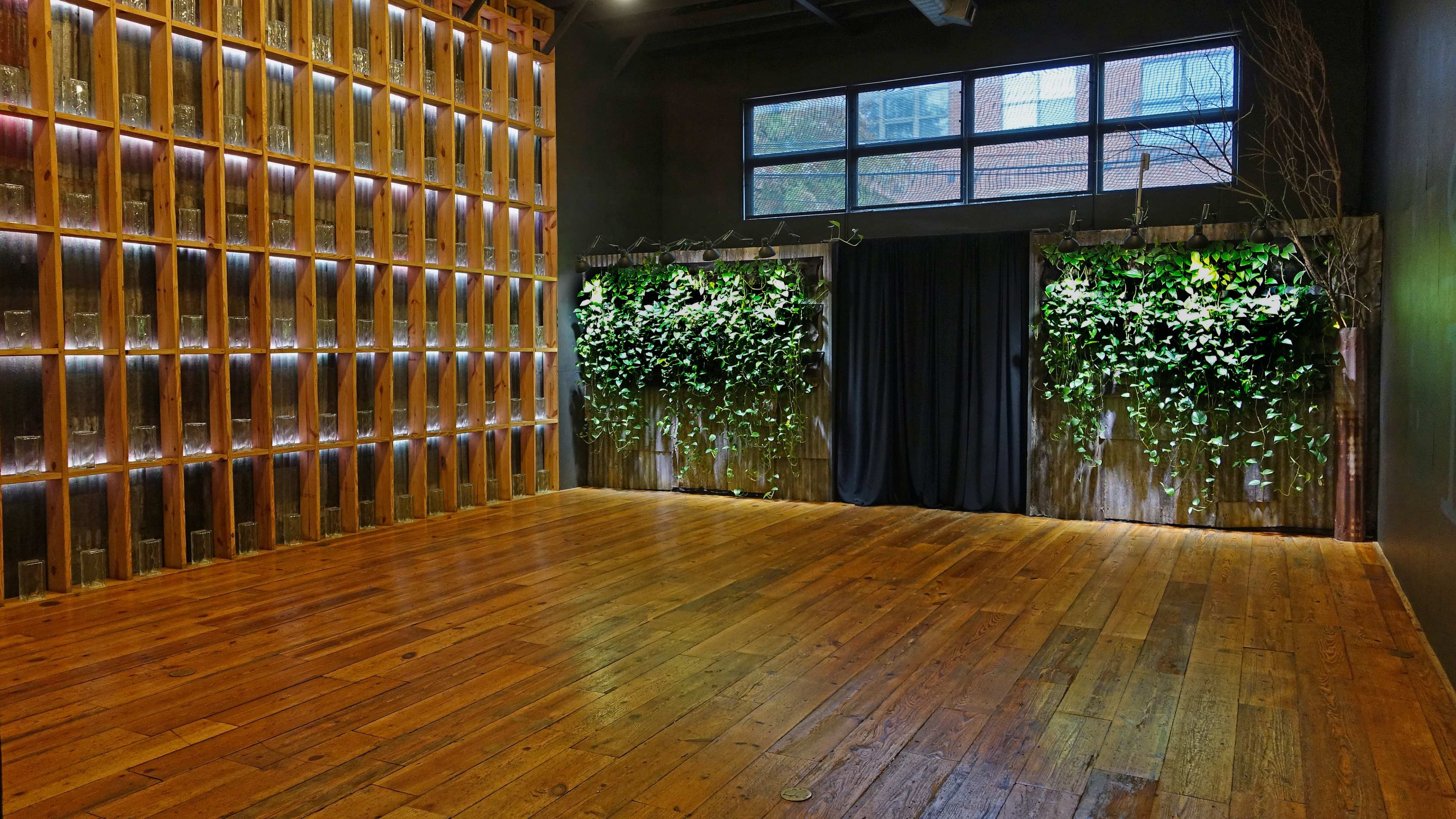 A spacious indoor venue with wooden flooring, a wall of lit glass containers, and greenery against a dark backdrop.