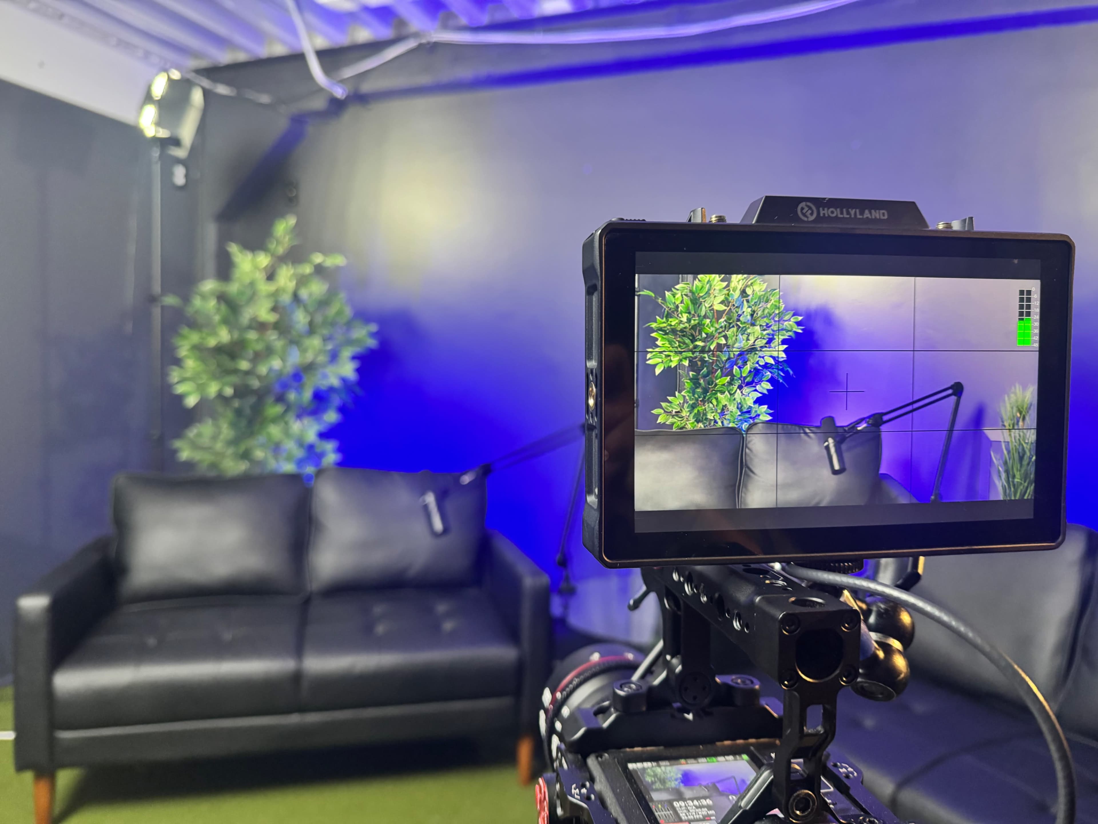 A camera viewfinder displays a black couch with plants in the background, set up in a studio environment.