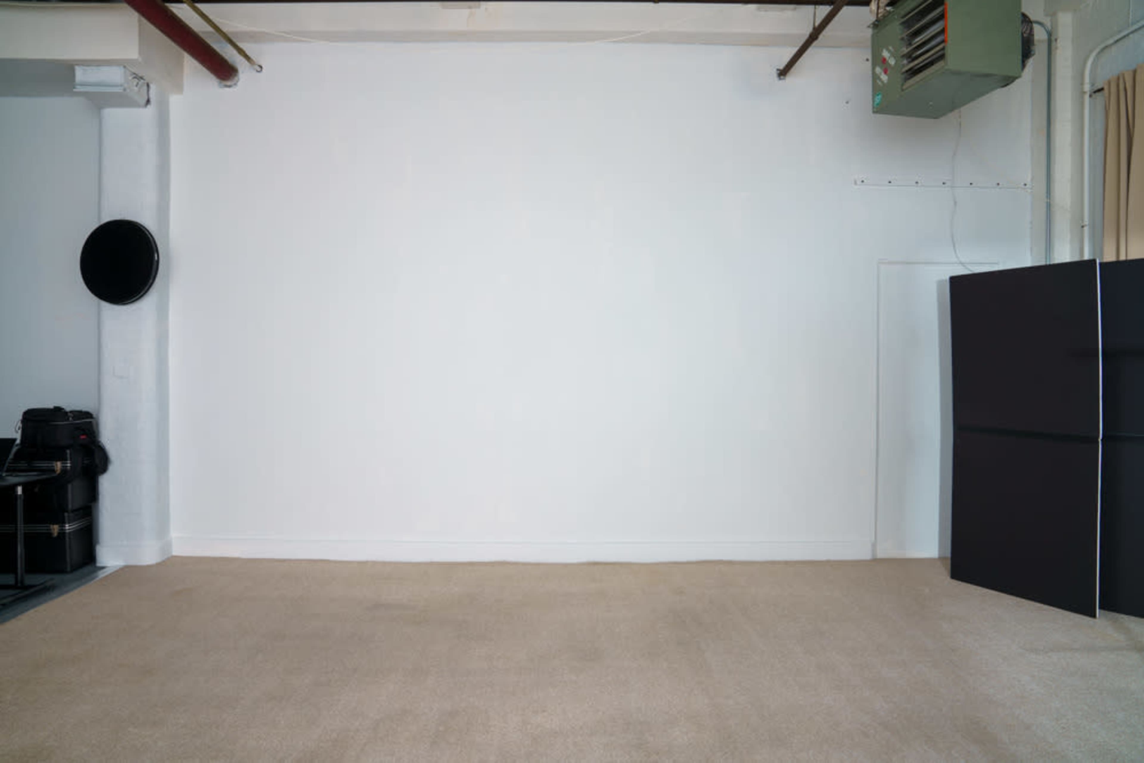 An empty room with a white wall, beige carpet, and a black refrigerator in the corner.