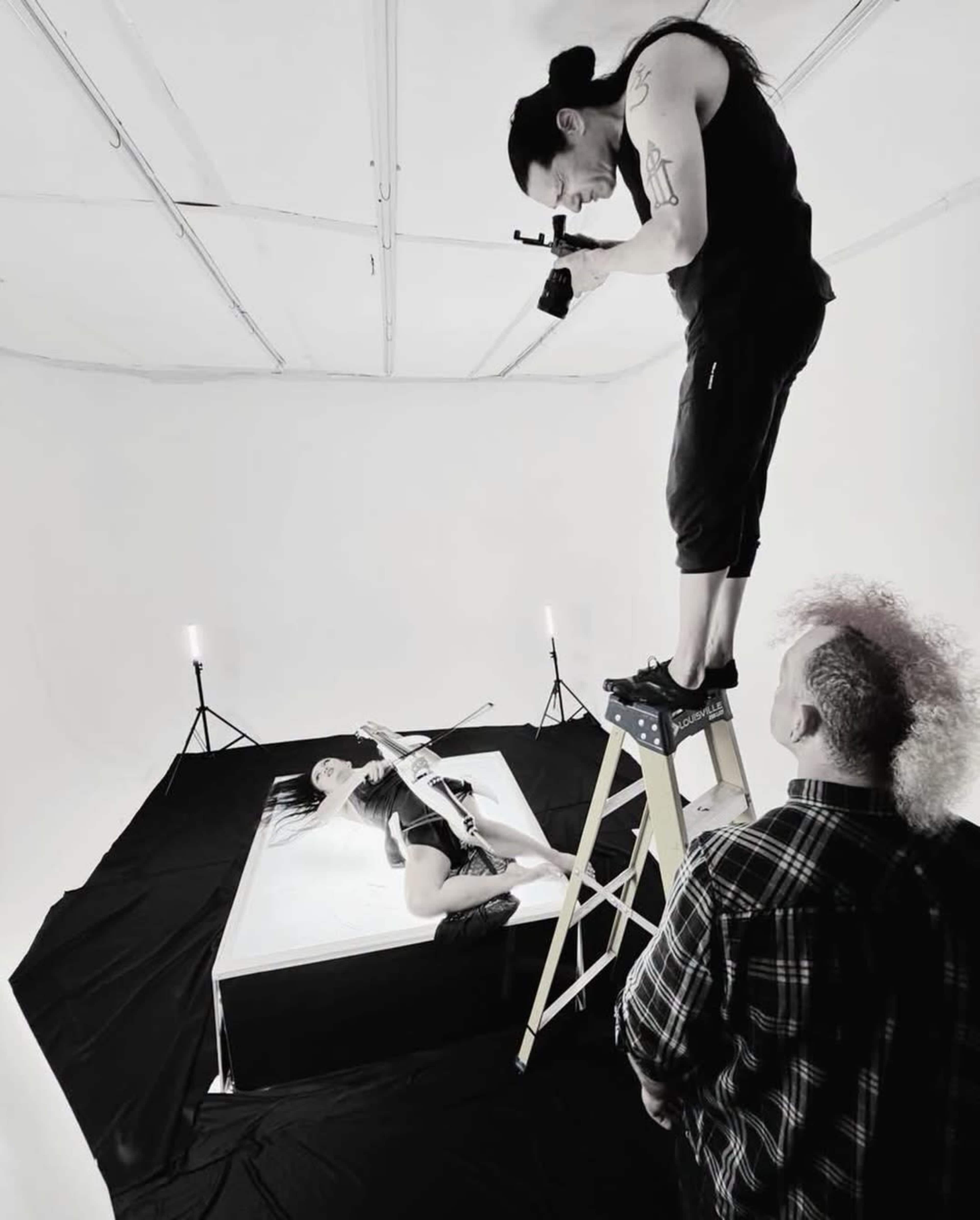 A photographer stands on a ladder above a model posed in a shallow tub, while another person observes from the ground in a stark white studio setting.