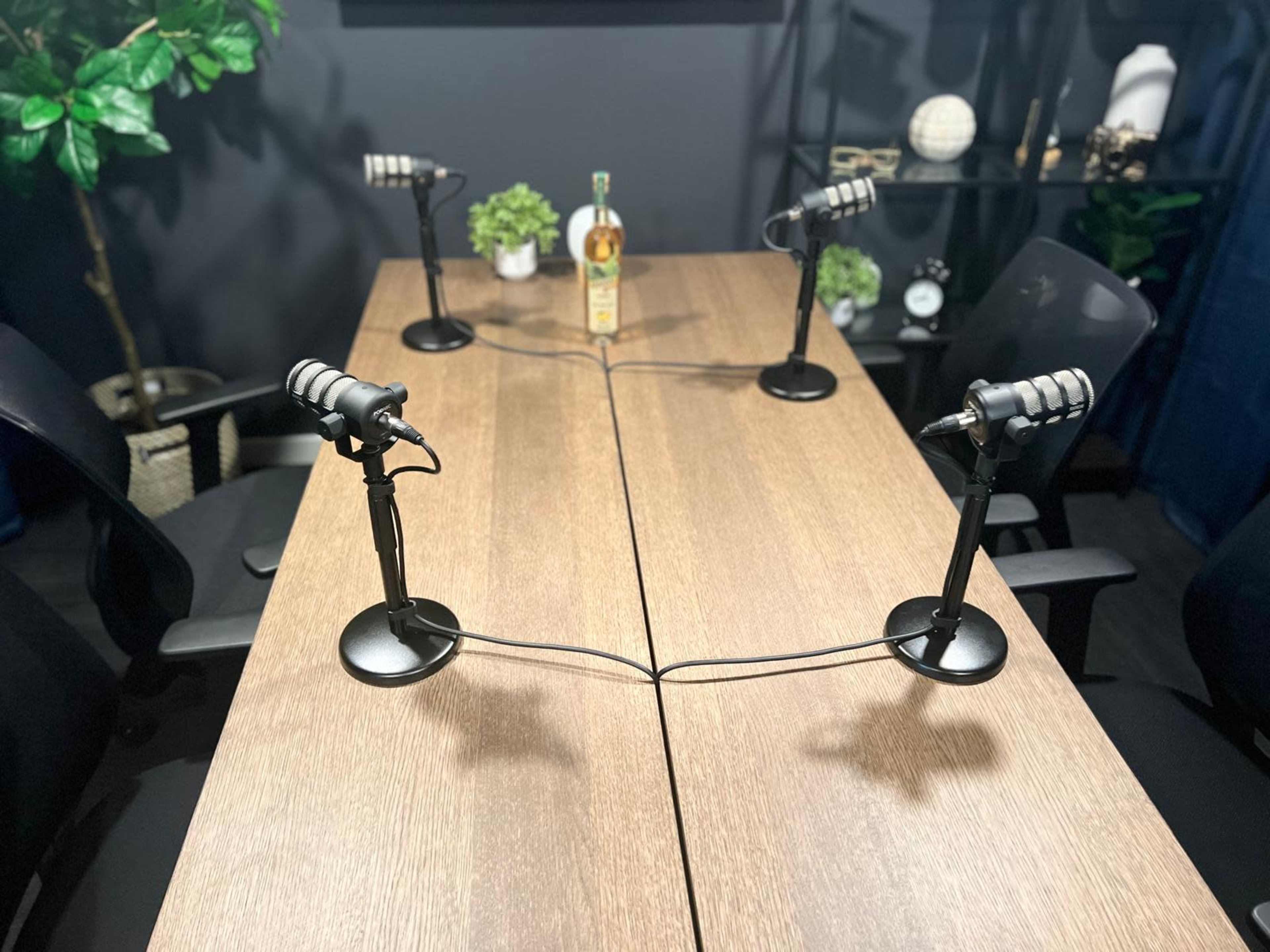 The image shows a podcasting setup with two microphones on stands positioned at a wooden table, surrounded by empty black chairs and some decorative items in the background.