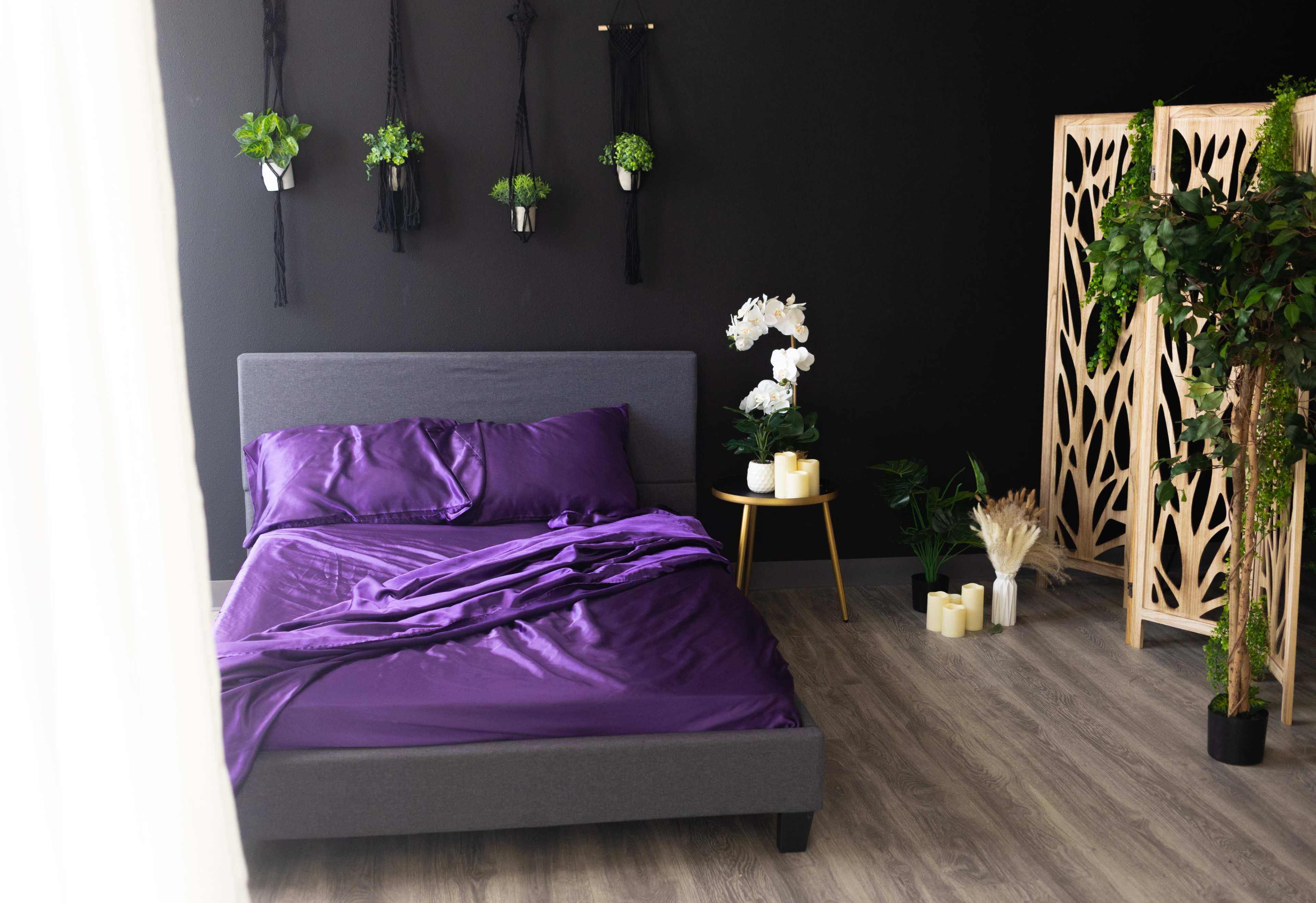 The image shows a modern bedroom featuring a gray bed with purple bedding, surrounded by plants and decorative screens.
