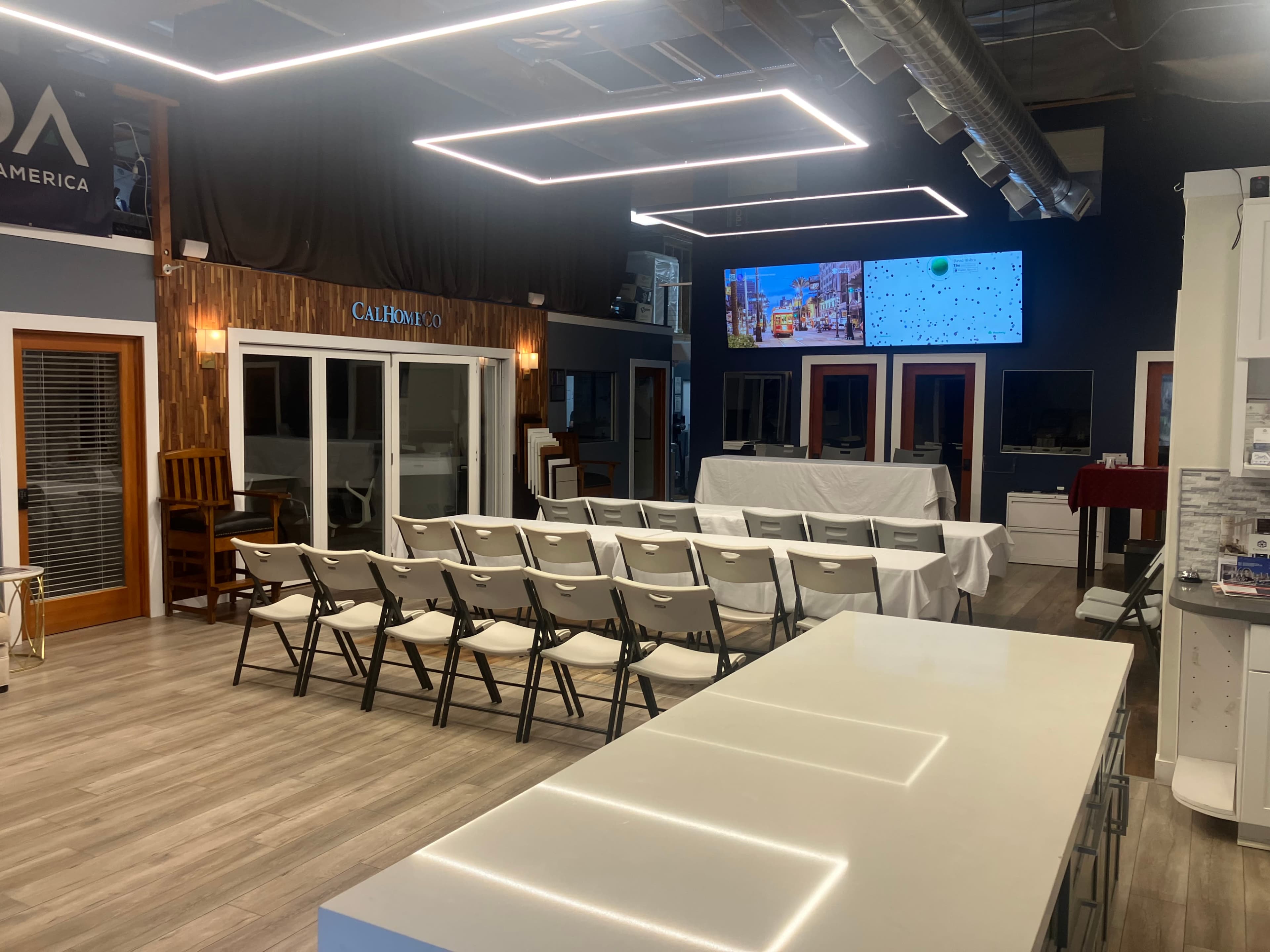 The image shows a spacious indoor area with rows of chairs facing a stage-like setup, a large screen on the wall, and various furniture displays.