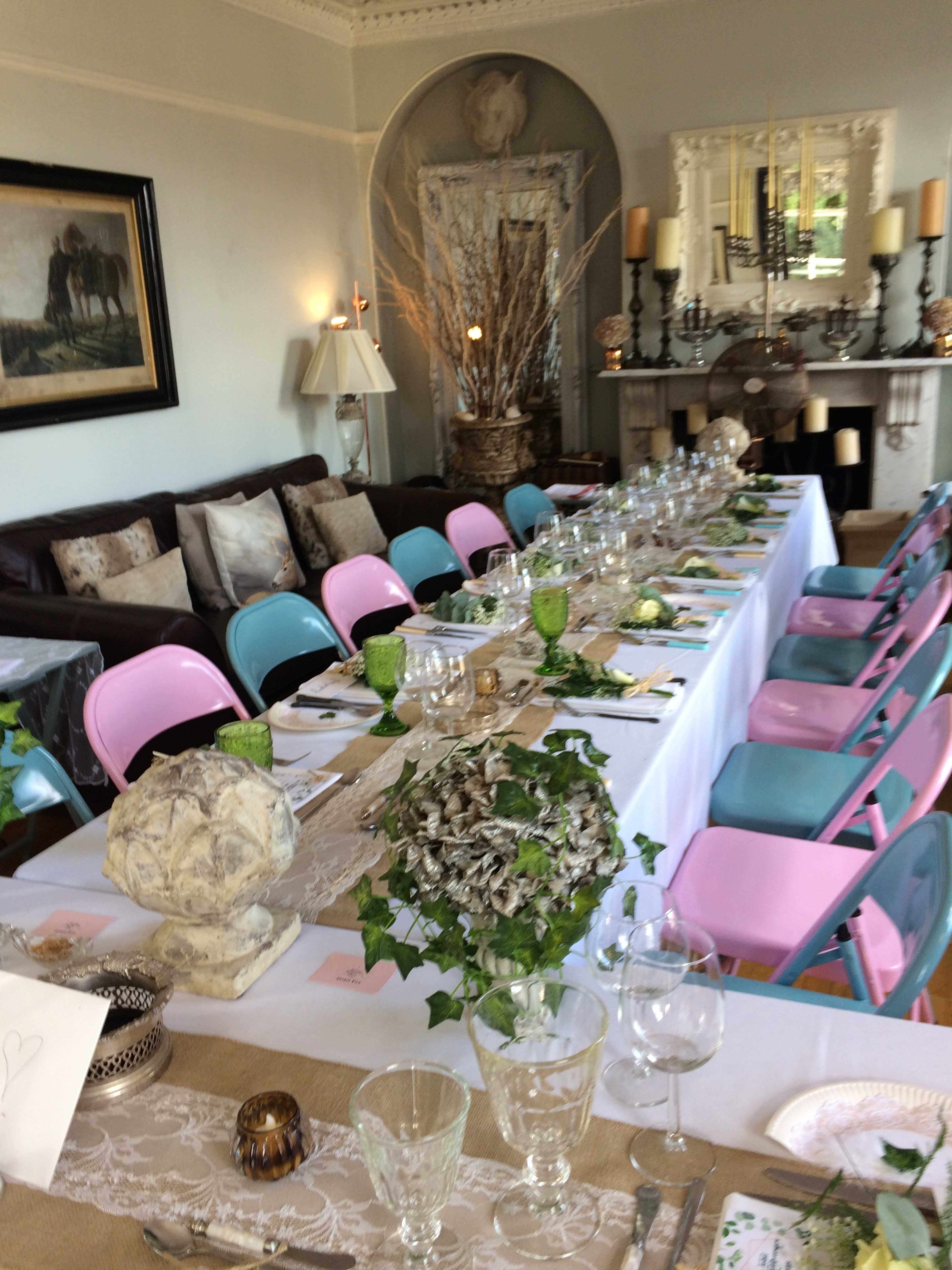 A long dining table set with glassware, cutlery, and decorative greenery is surrounded by chairs in pink and blue, with a stylish living room in the background.