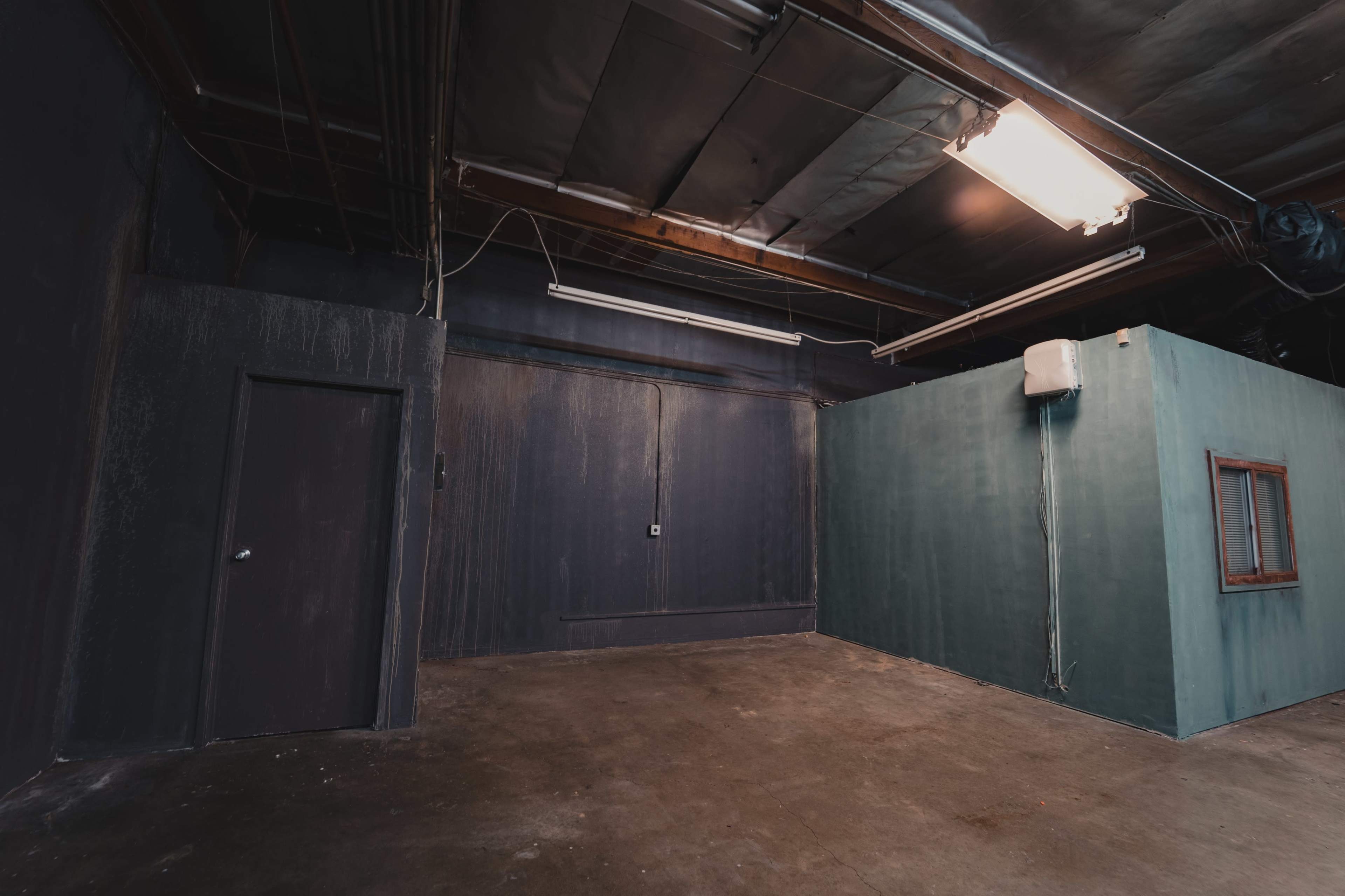 An industrial room with one dark wall, a blue-green wall with a small window, and a door on the left side.