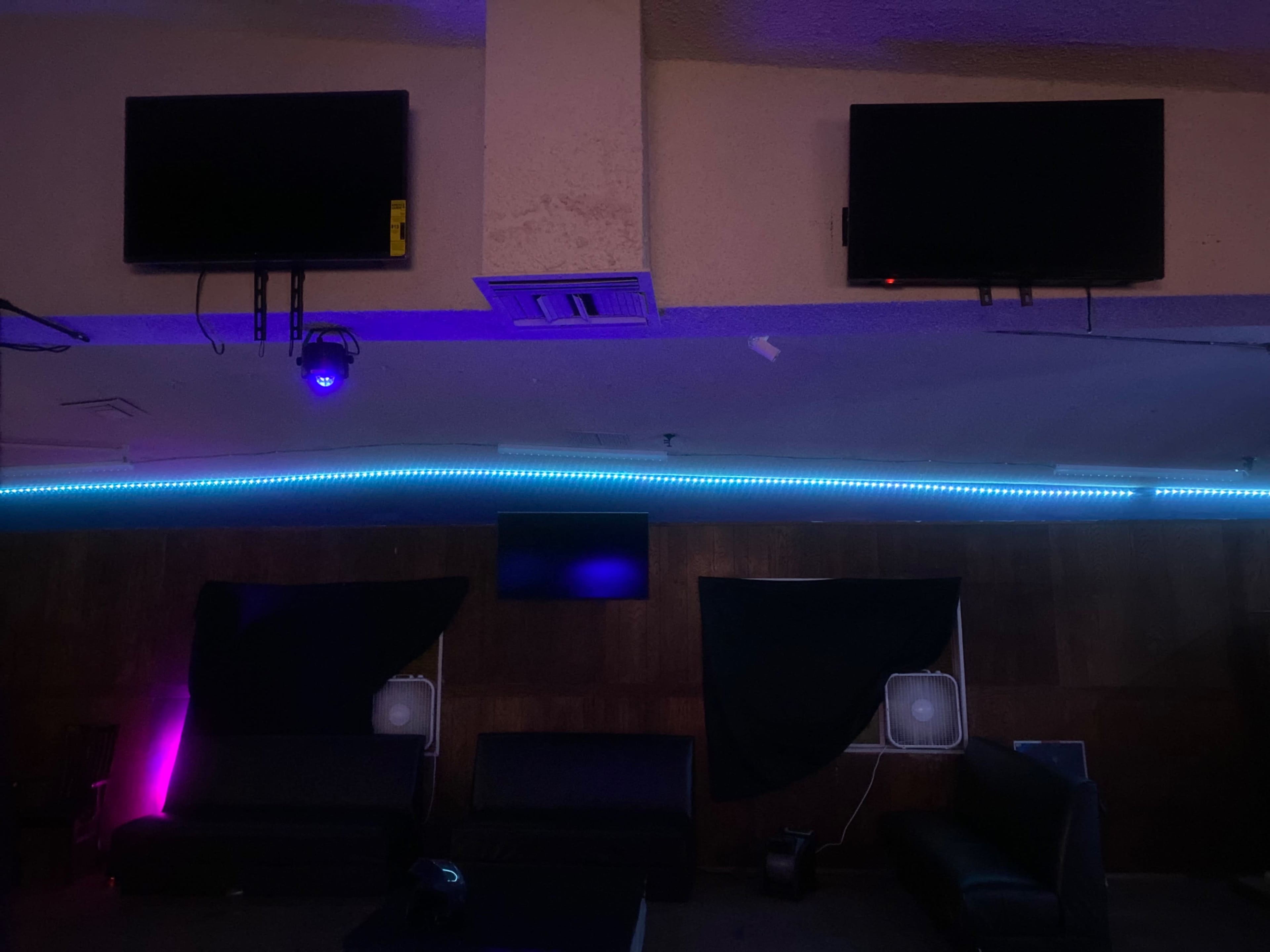 A dimly lit room with two televisions mounted on the wall, a blue LED light strip along the lower edge, and dark drapes covering some areas.