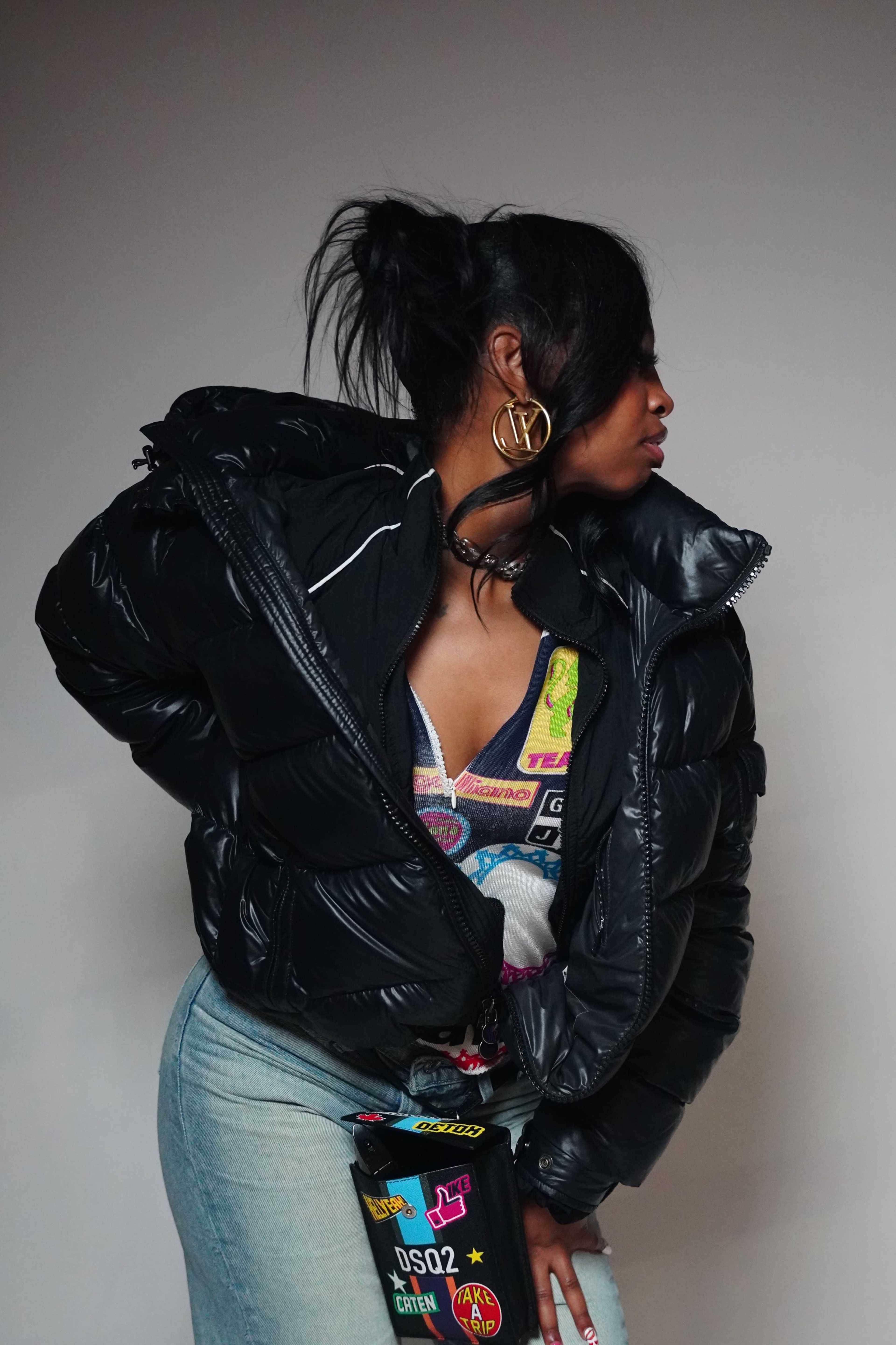 A person is wearing a large black puffer jacket and standing in profile, with their hair styled up and a colorful graphic top underneath.