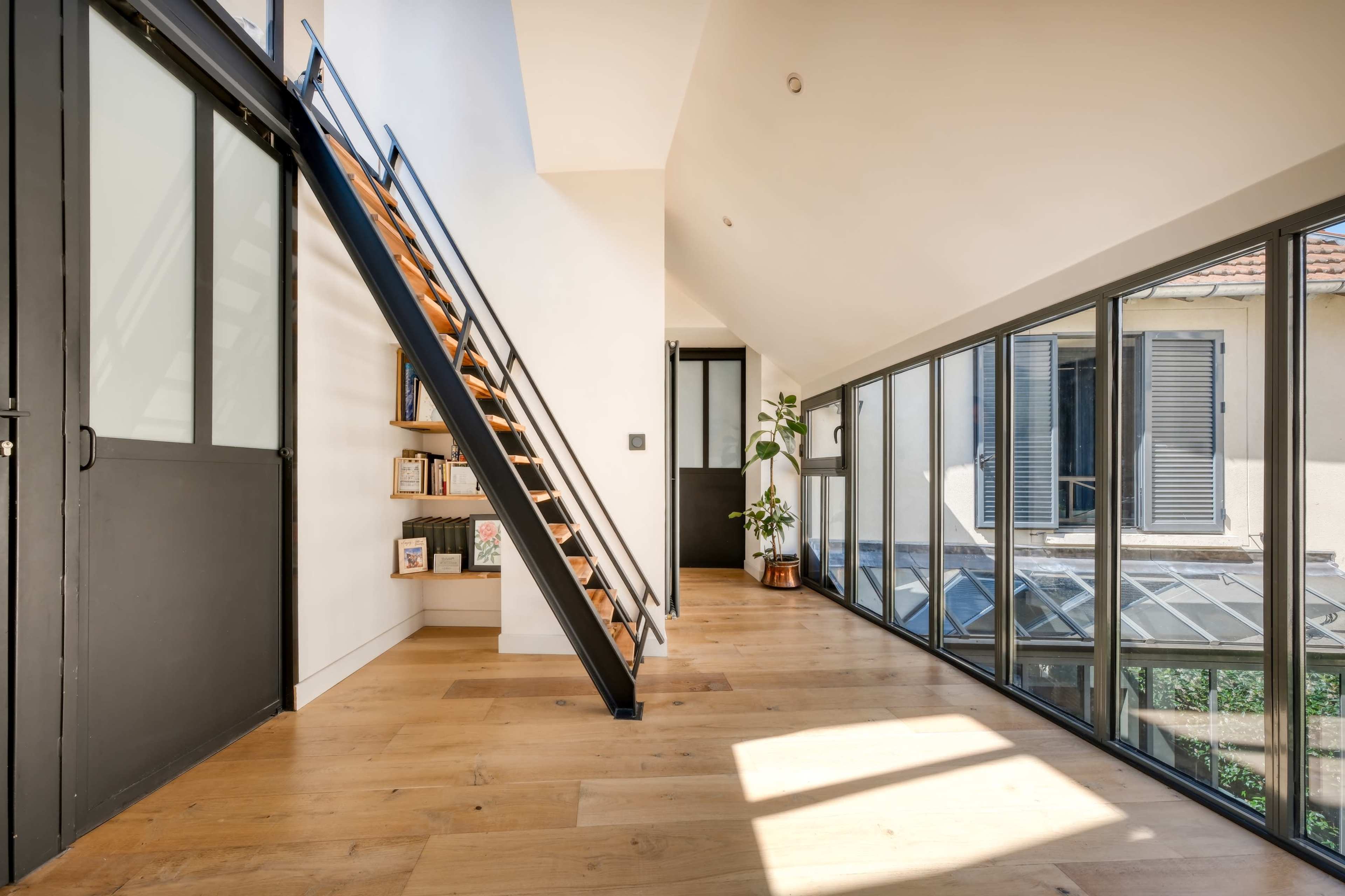 The image features a modern interior space with a staircase leading to an upper level, large windows allowing natural light, and a minimalist design.