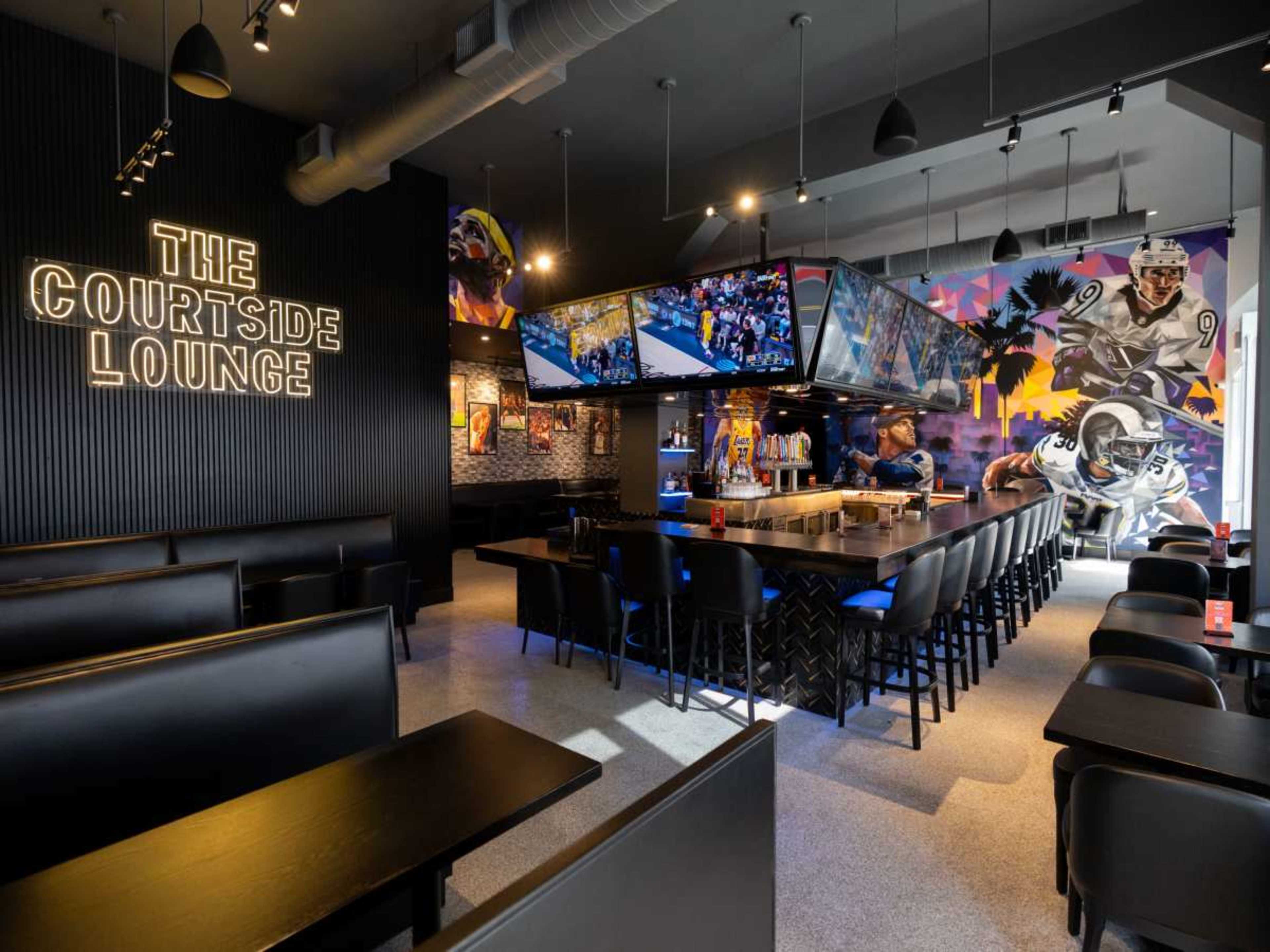 The image shows a modern lounge area with a bar, multiple large screens displaying sports, and vibrant murals on the walls.