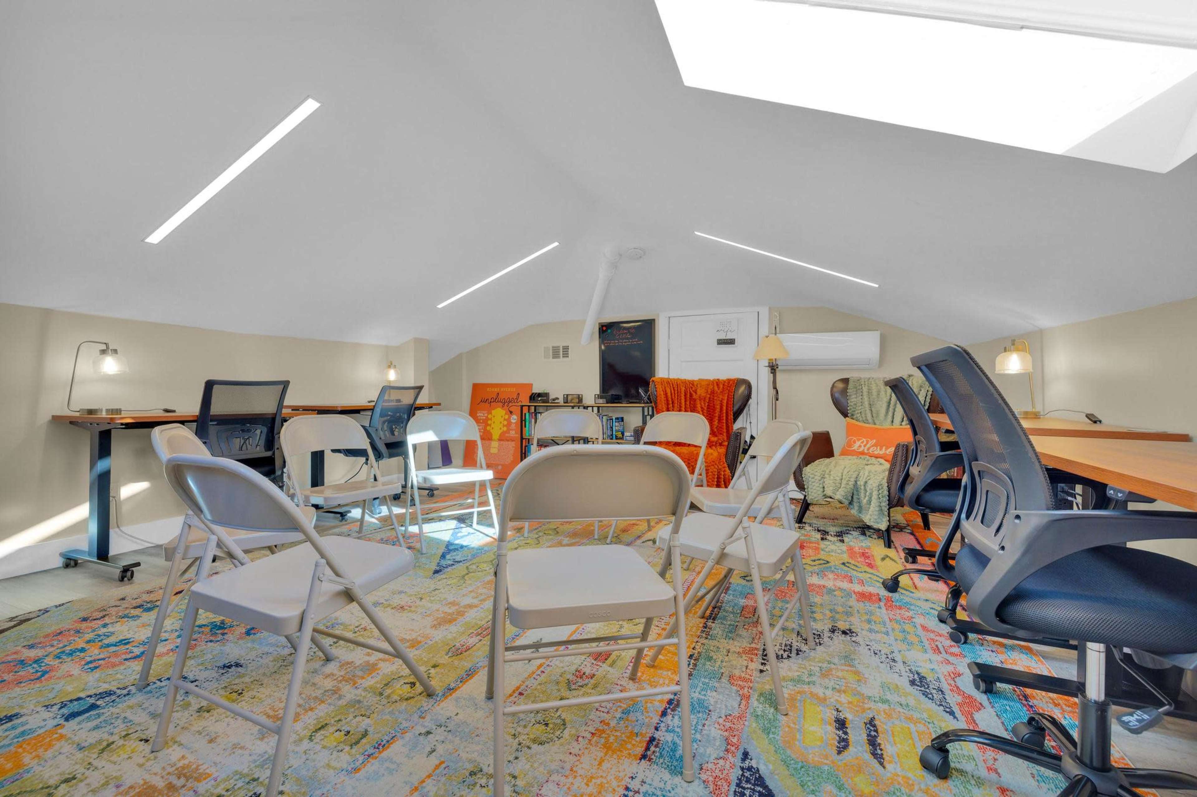 An attic room arranged with folding chairs in a circle on a colorful rug, surrounded by desks and computer setups.