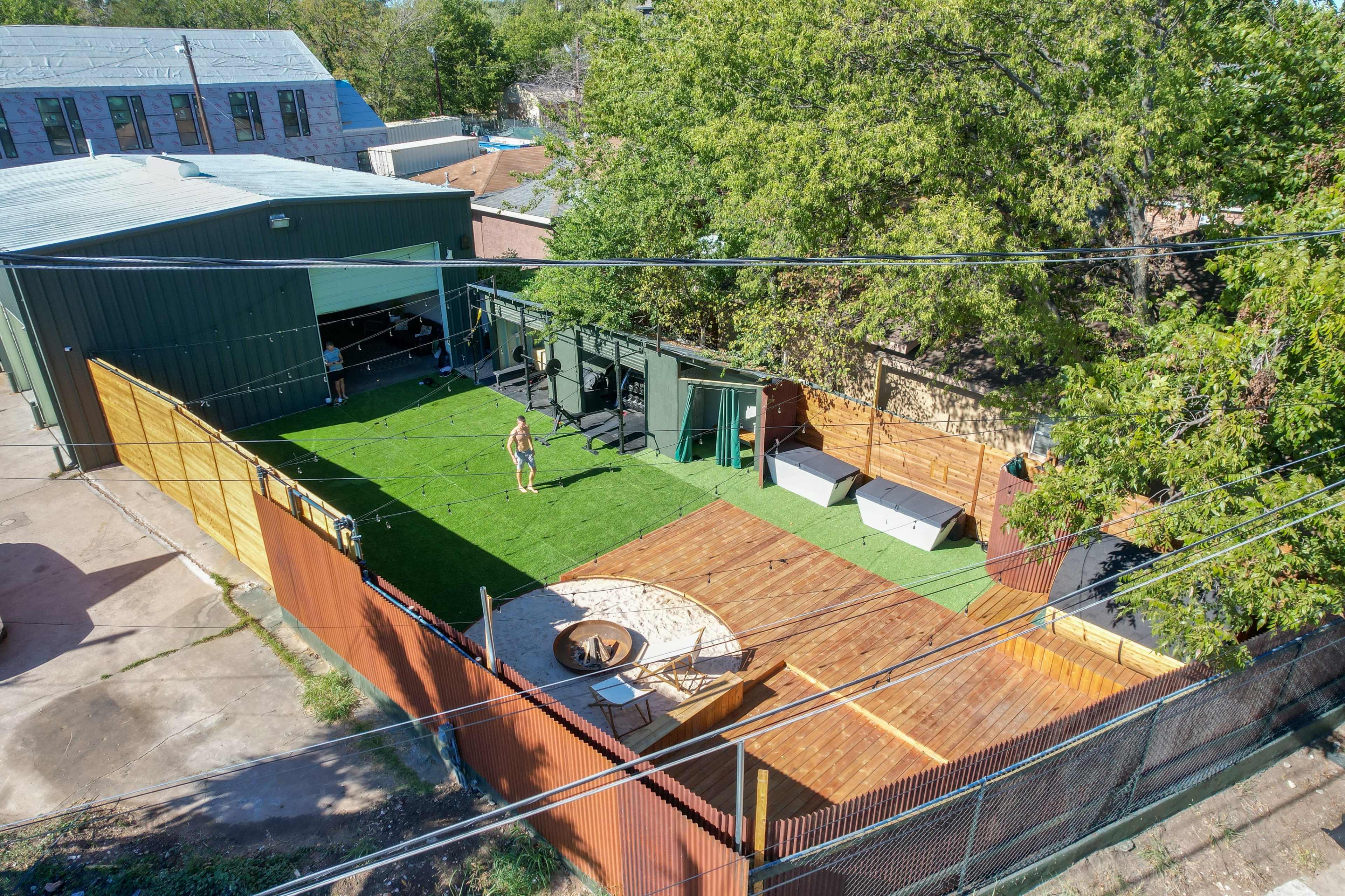 The image shows an outdoor space featuring green grass, wooden decks, seating areas, and a circular fire pit, surrounded by fences and buildings.