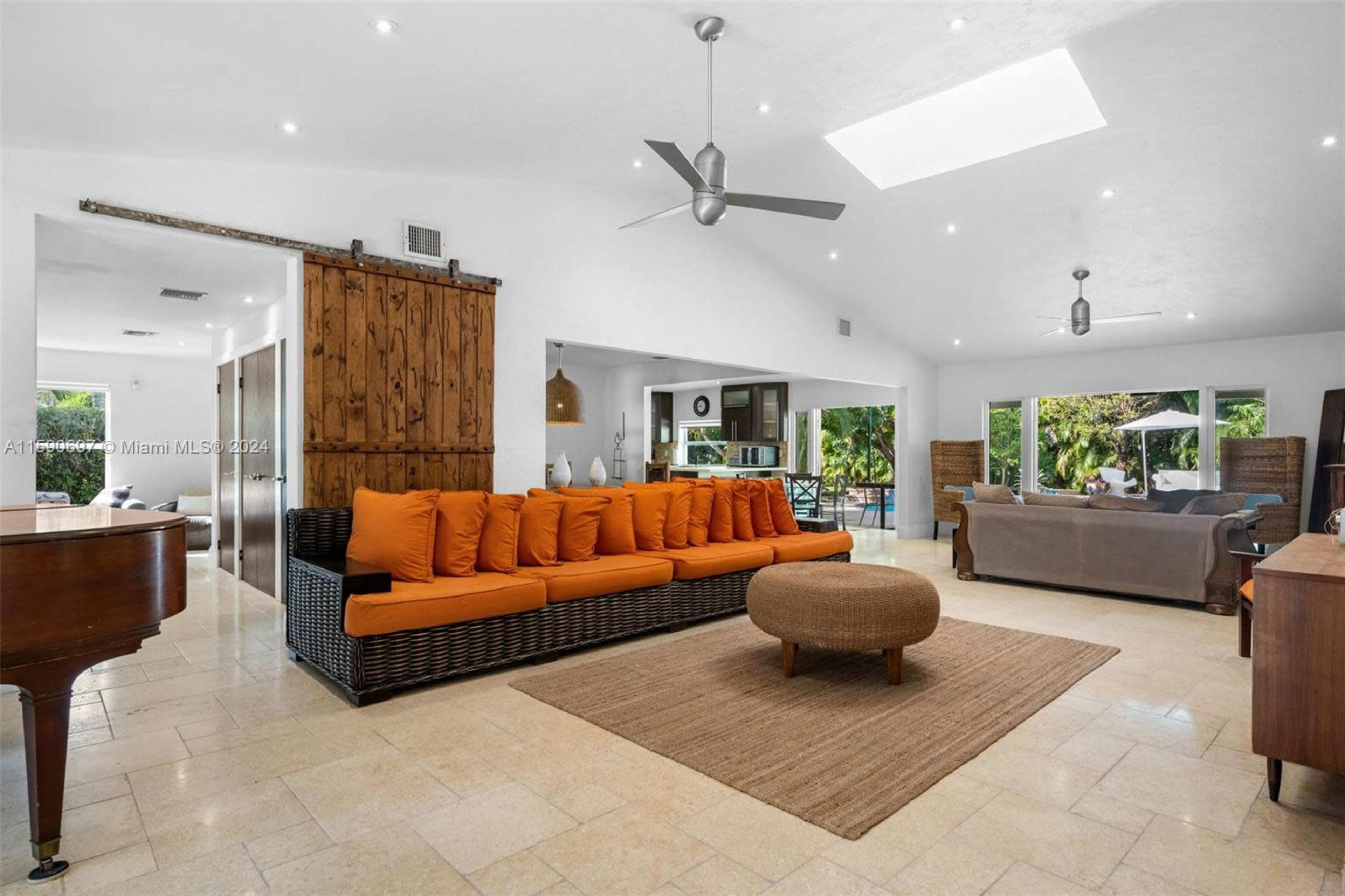 The image shows a spacious living area featuring a large orange sofa, a round coffee table, and a grand piano, with large windows overlooking a garden.