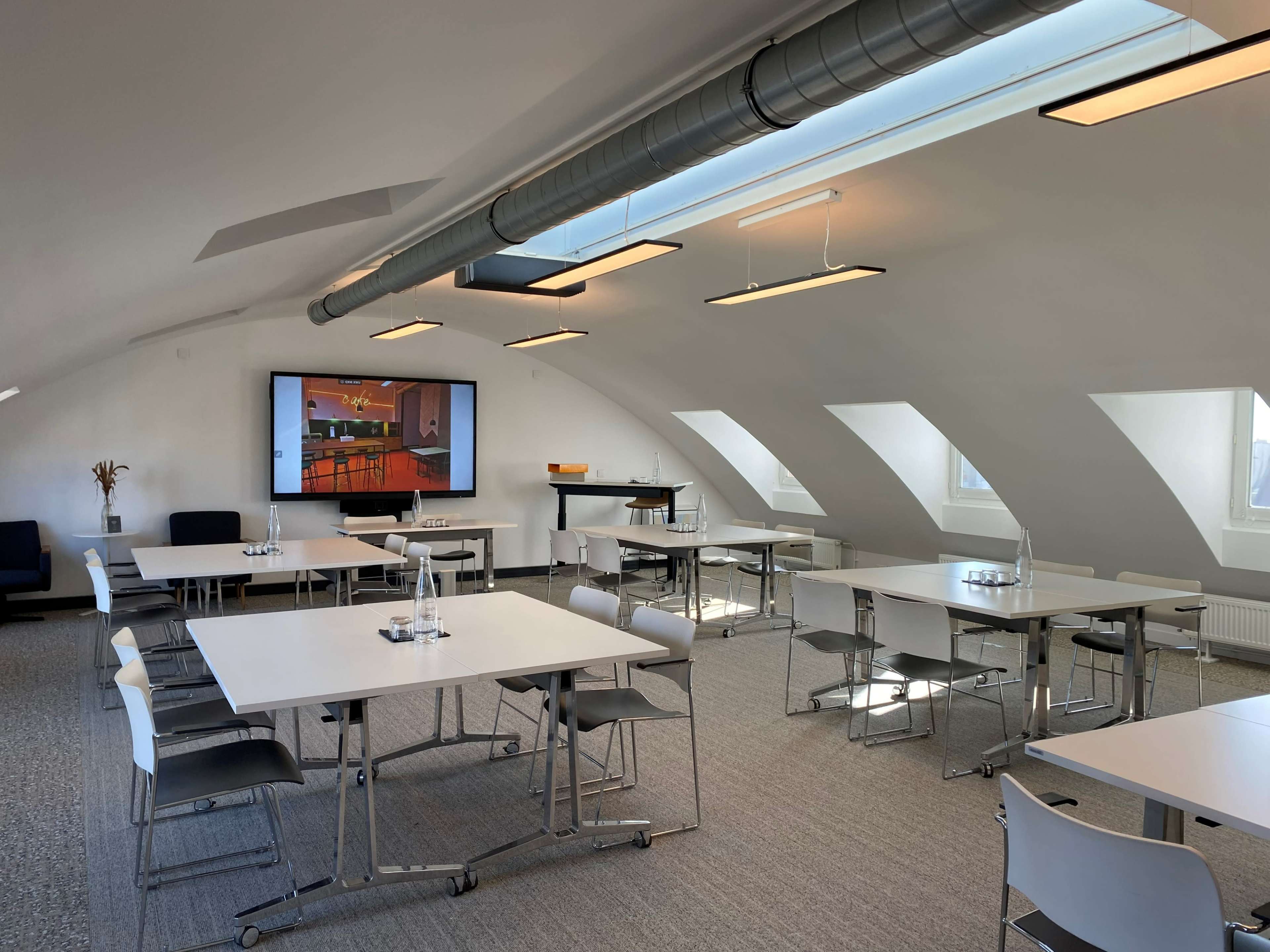A modern meeting room features multiple tables and chairs arranged around a central area with a large screen displaying a presentation.