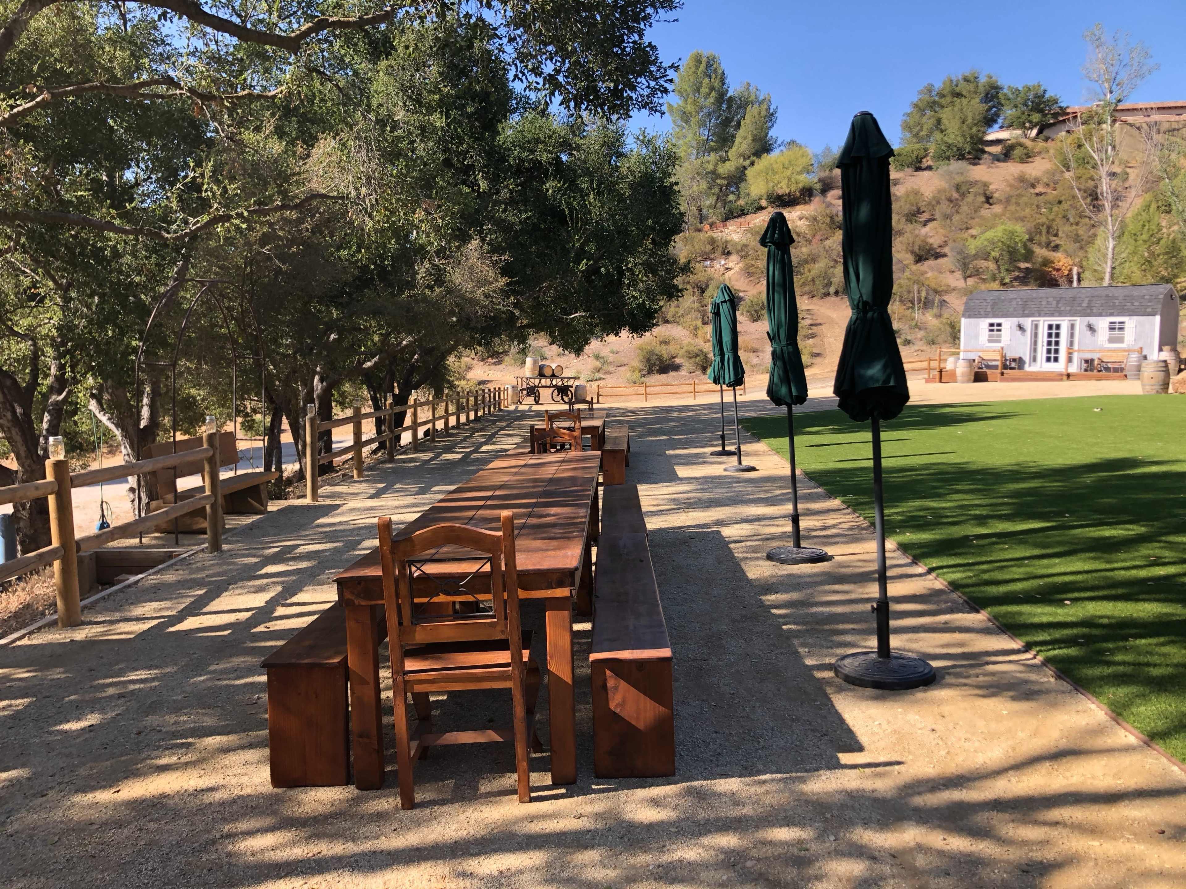 A long wooden table with chairs is set up under green umbrellas alongside a grassy area and rustic buildings in a rural setting.