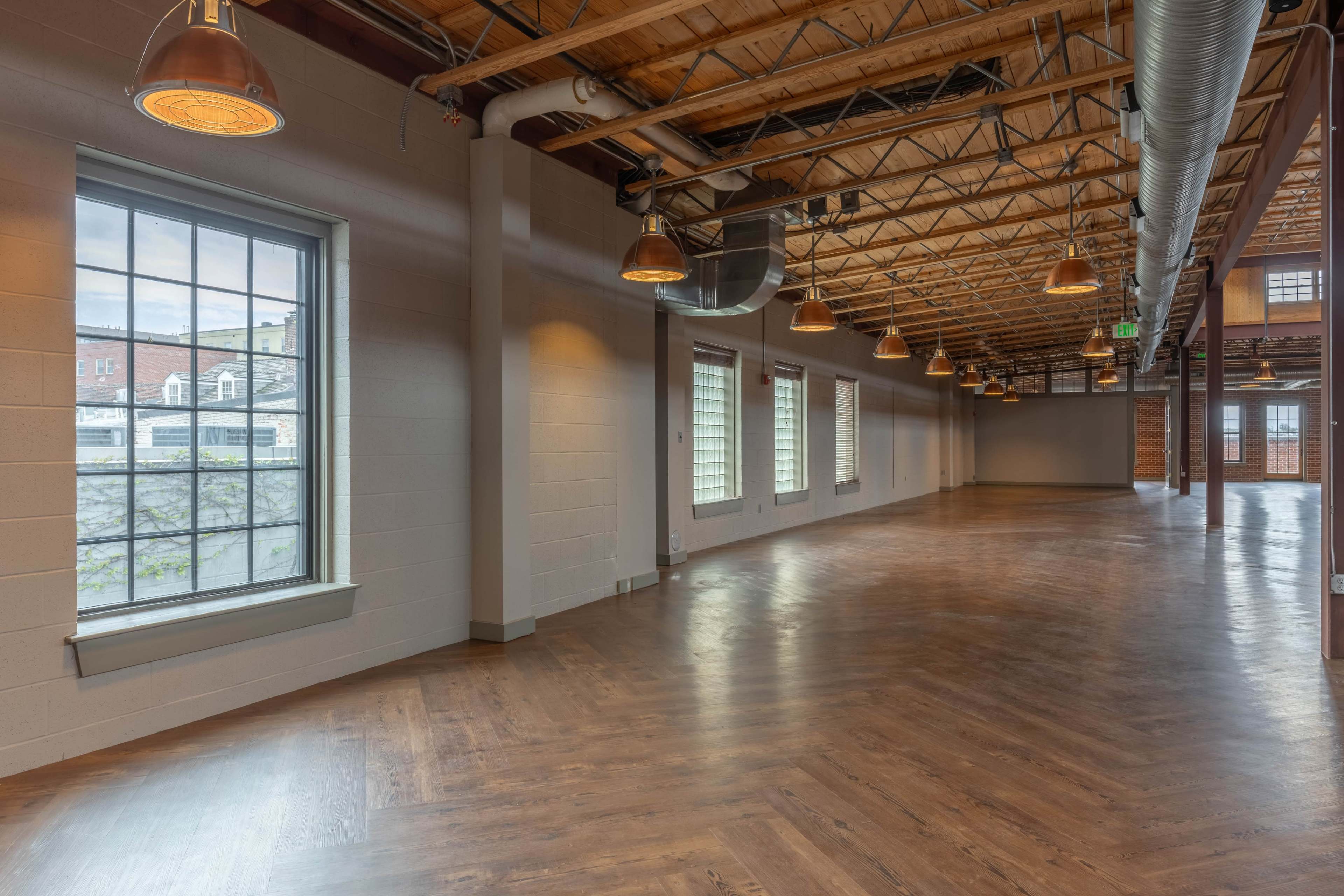 The image shows an empty room with polished wooden flooring, large windows, and exposed wooden beams in the ceiling, illuminated by industrial light fixtures.