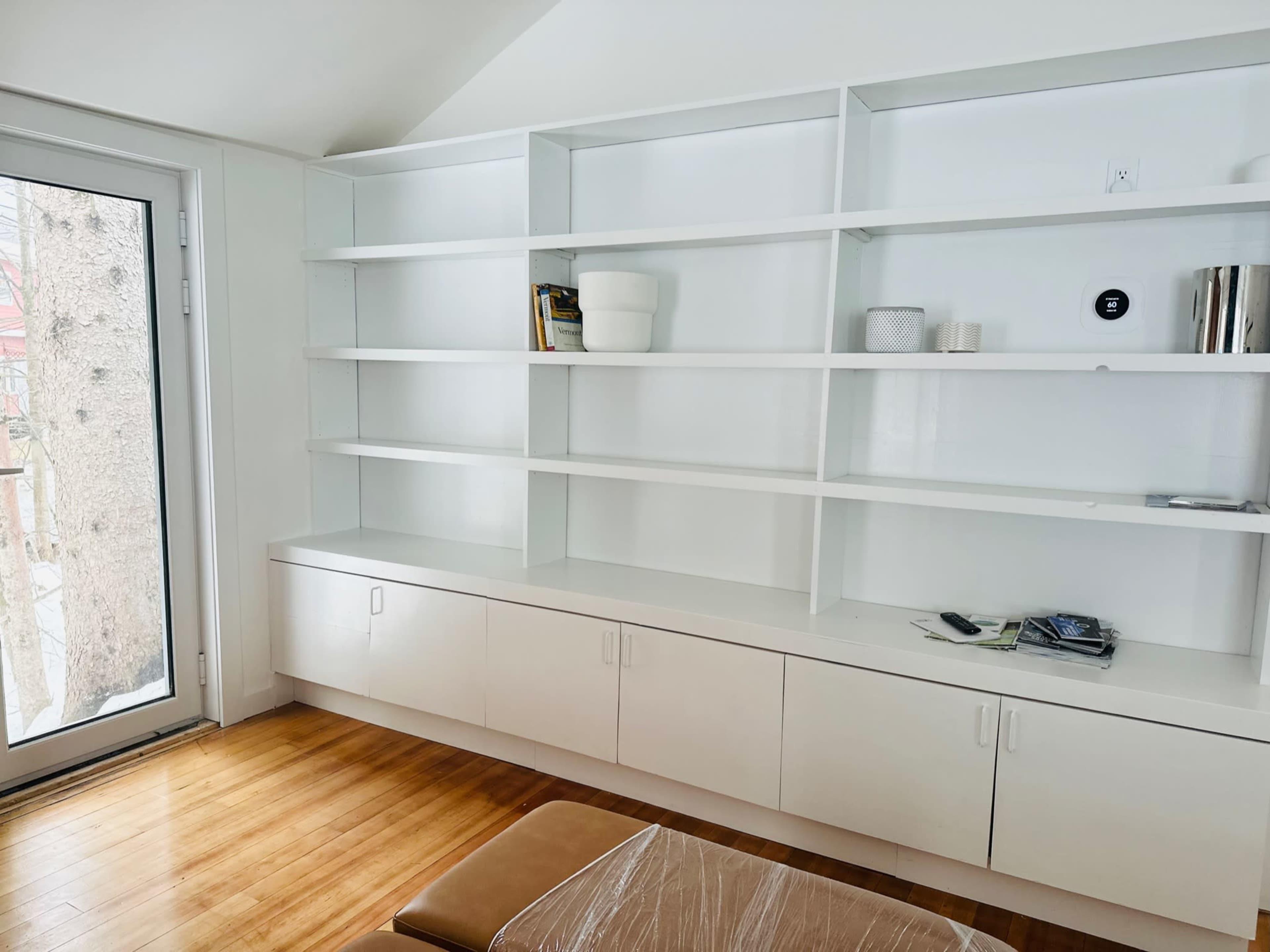 A room features a white shelving unit and cabinets, with a view of a tree through a glass door.