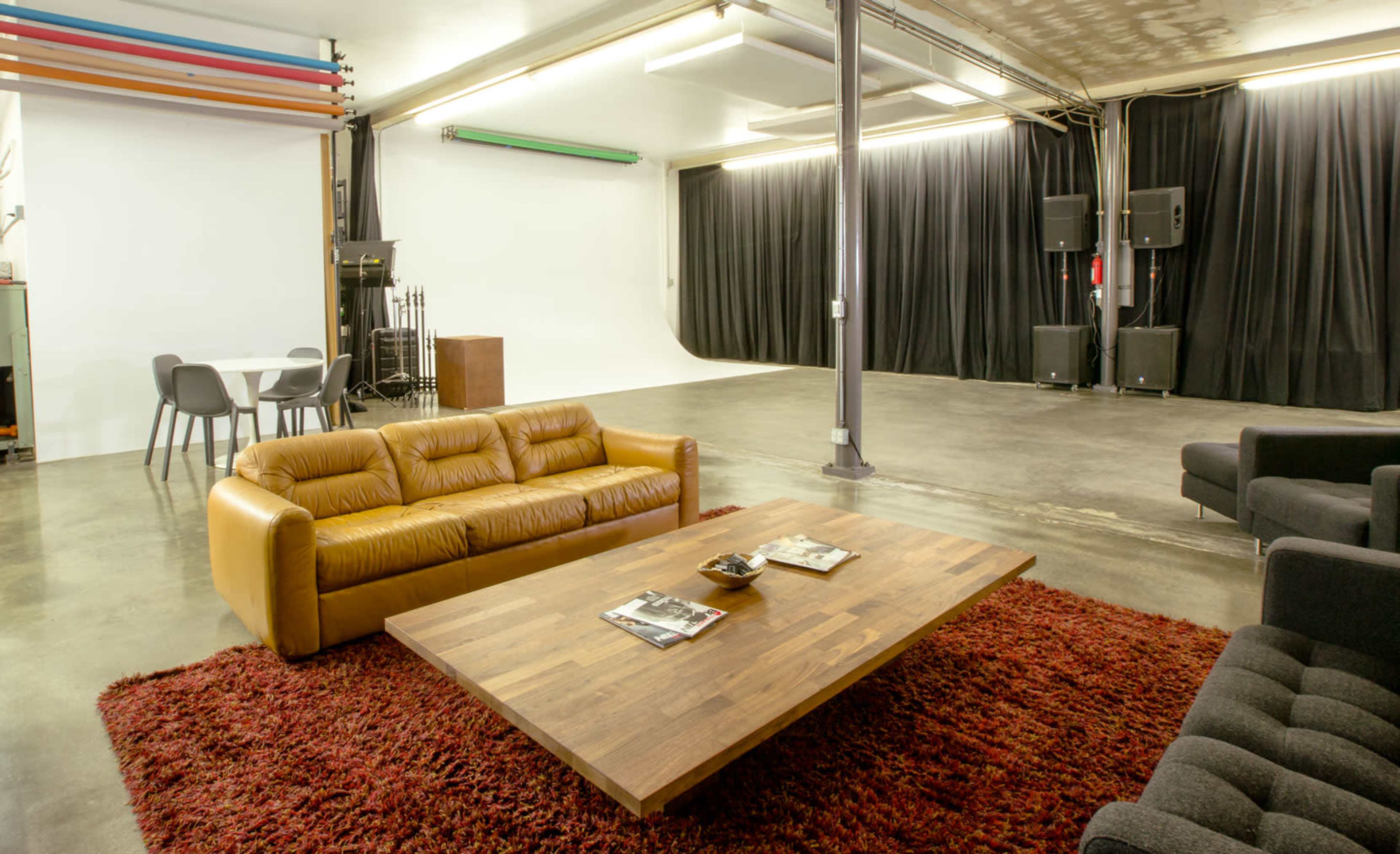 The image shows a spacious studio with a large wooden table, tan leather sofa, dark upholstered couch, and a backdrop of black curtains and lighting equipment.