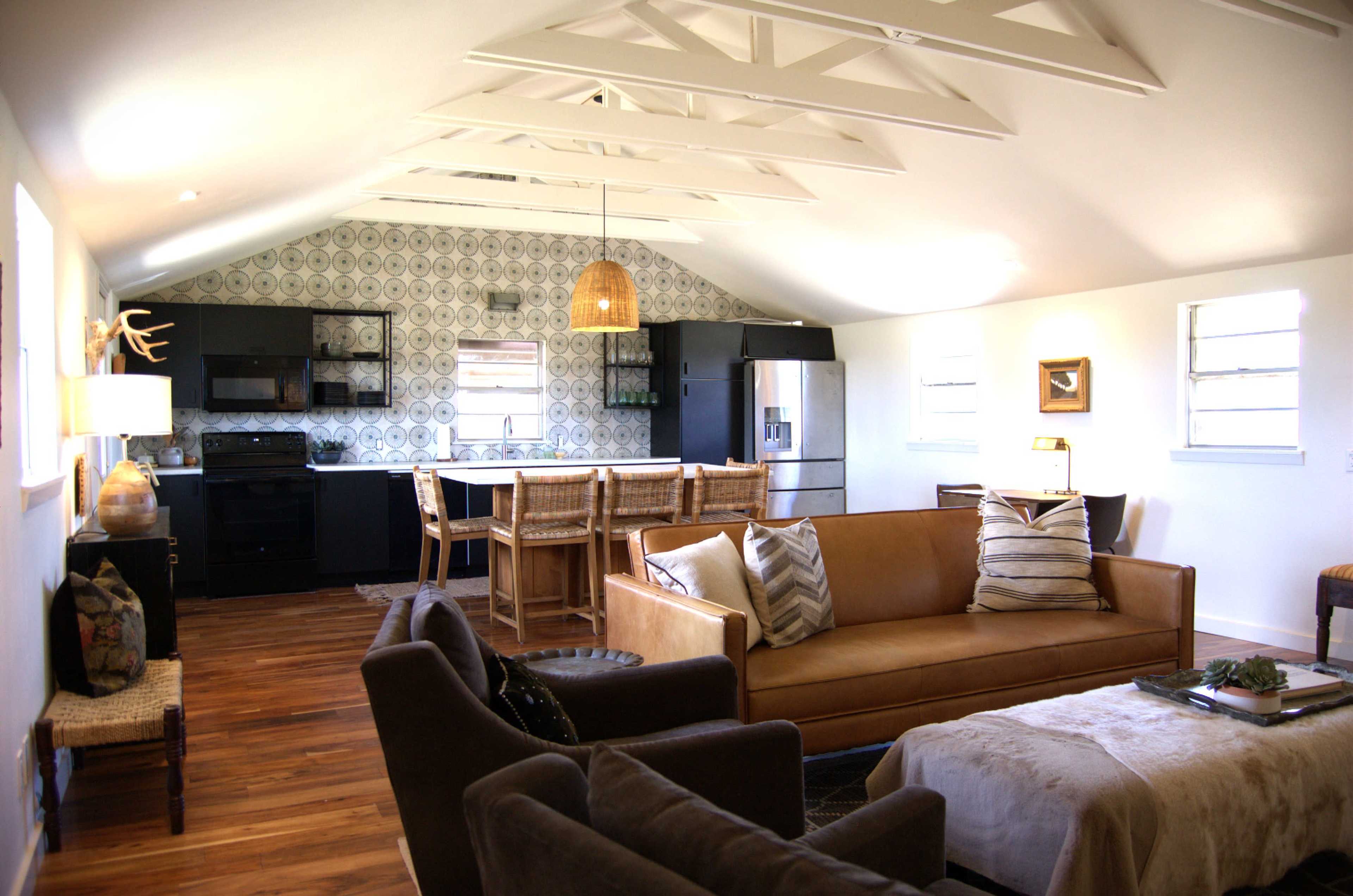 A modern, open-concept living space features a brown leather sofa, a gray chair, and a kitchen with black cabinets and a patterned backsplash.