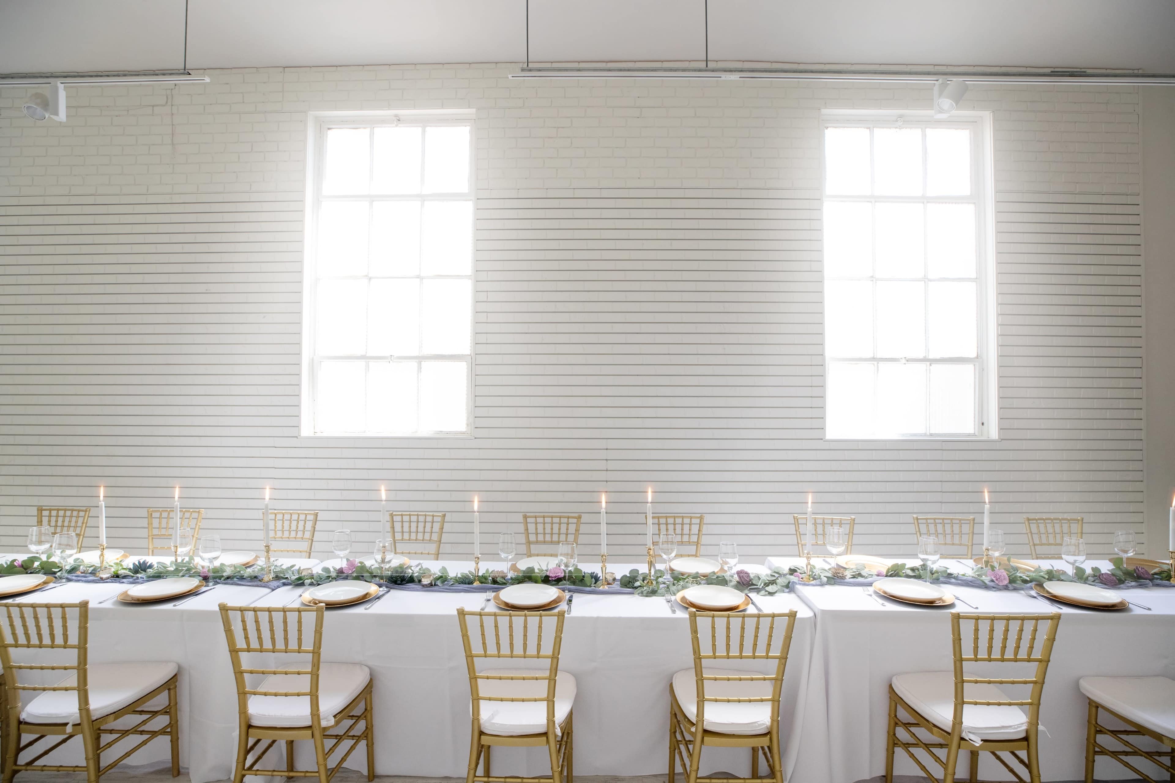 A long banquet table with white linens and gold chairs is set up against a wall with large windows, featuring a centerpiece of greenery and candles.