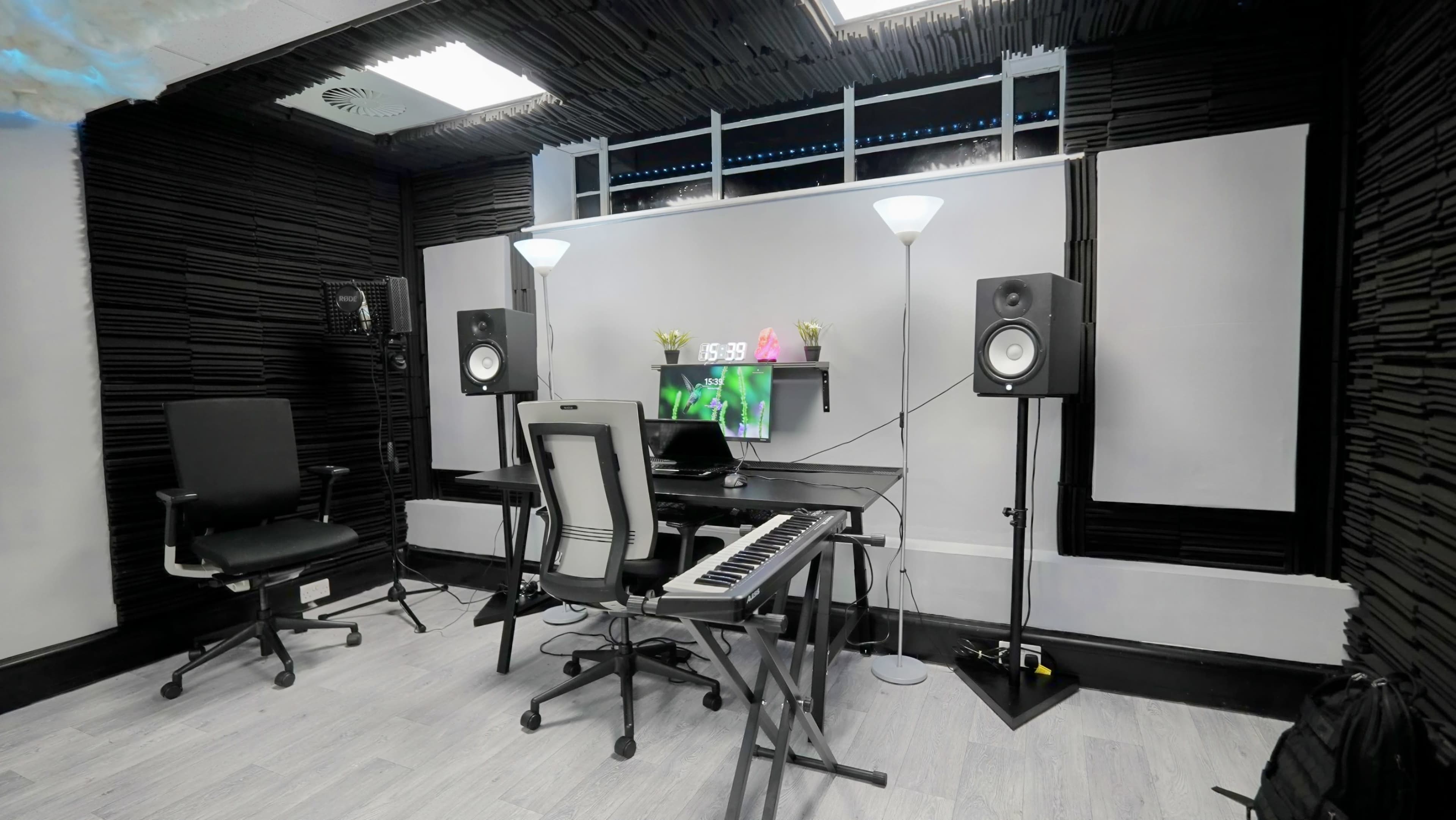 A modern music production studio featuring a desk with a computer, a keyboard, two studio monitors, and acoustic panels on the walls.