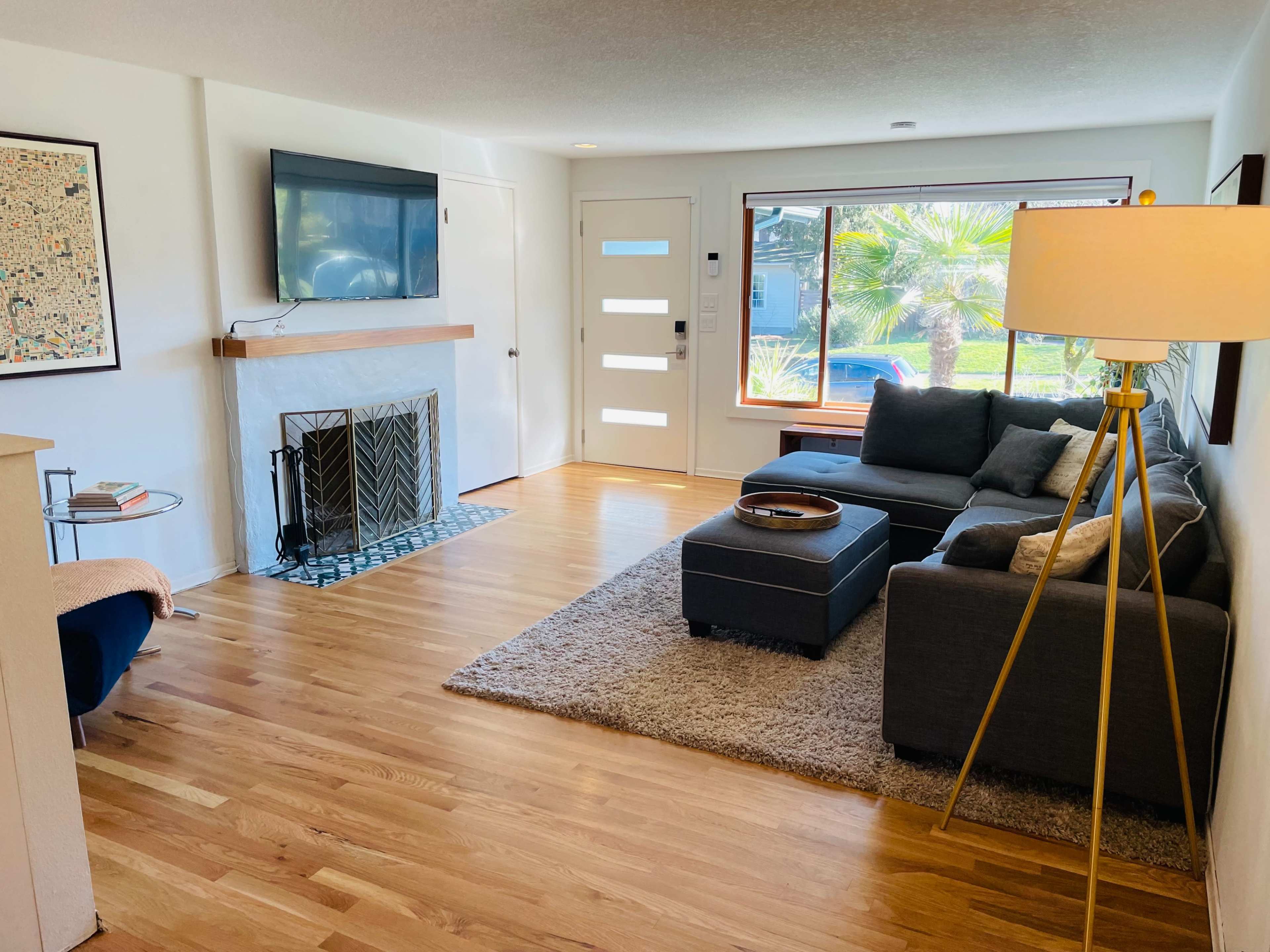 The living room features a gray sectional sofa, a coffee table, a flat-screen TV mounted on the wall, and a fireplace, with large windows overlooking a garden.