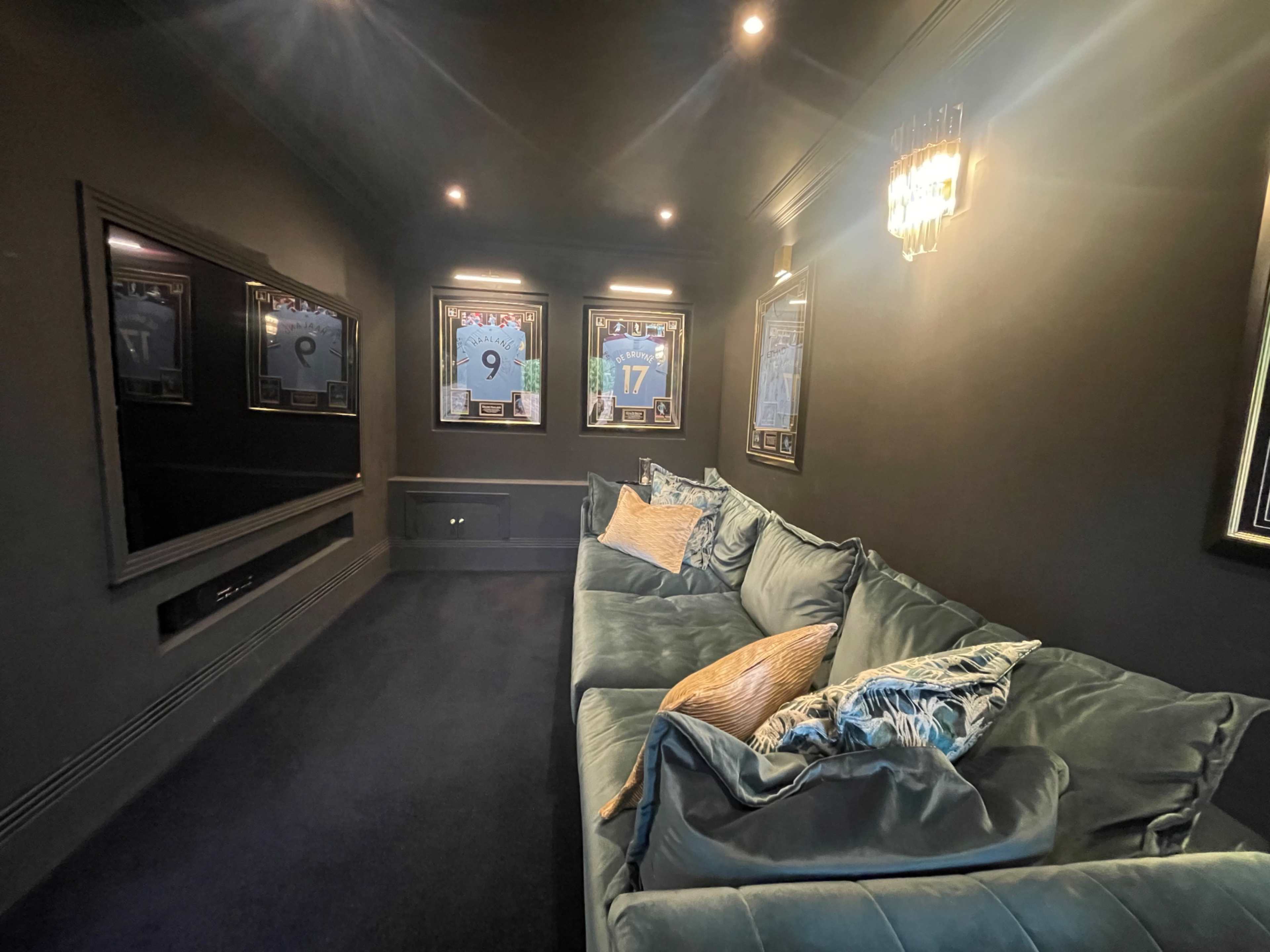 The image shows a darkened home theater with a plush sofa, framed sports jerseys on the walls, and decorative lighting fixtures.