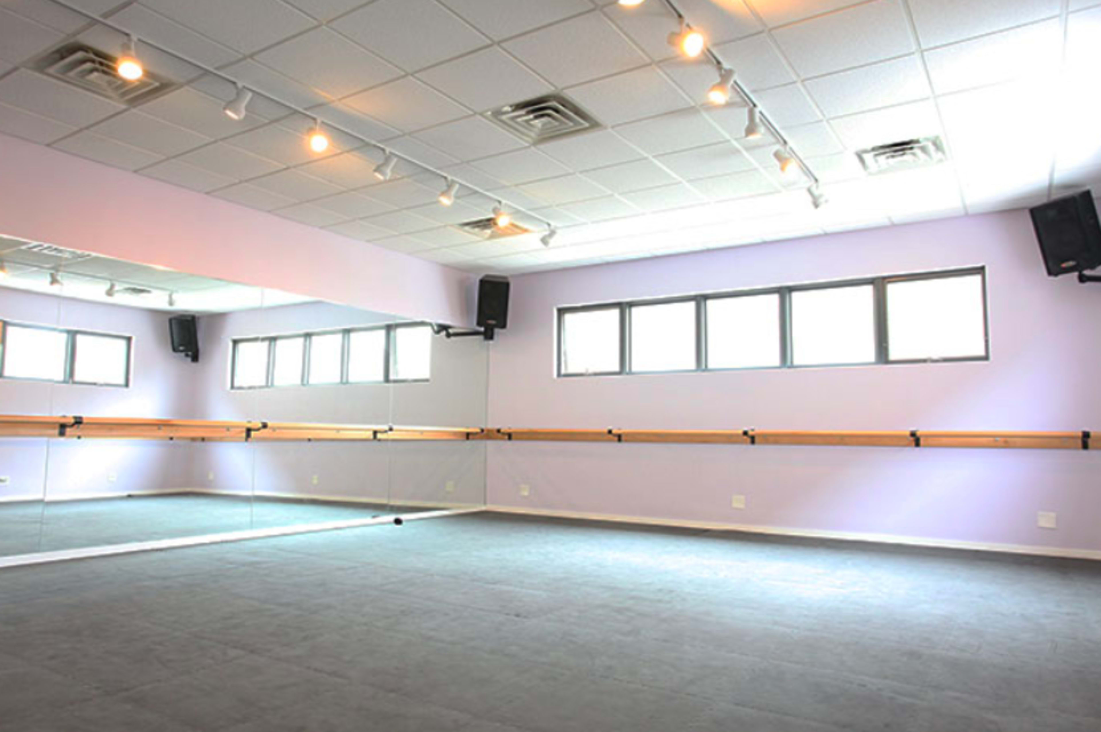 The image shows a dance studio with a mirrored wall, barre, and soft gray flooring under bright overhead lights.