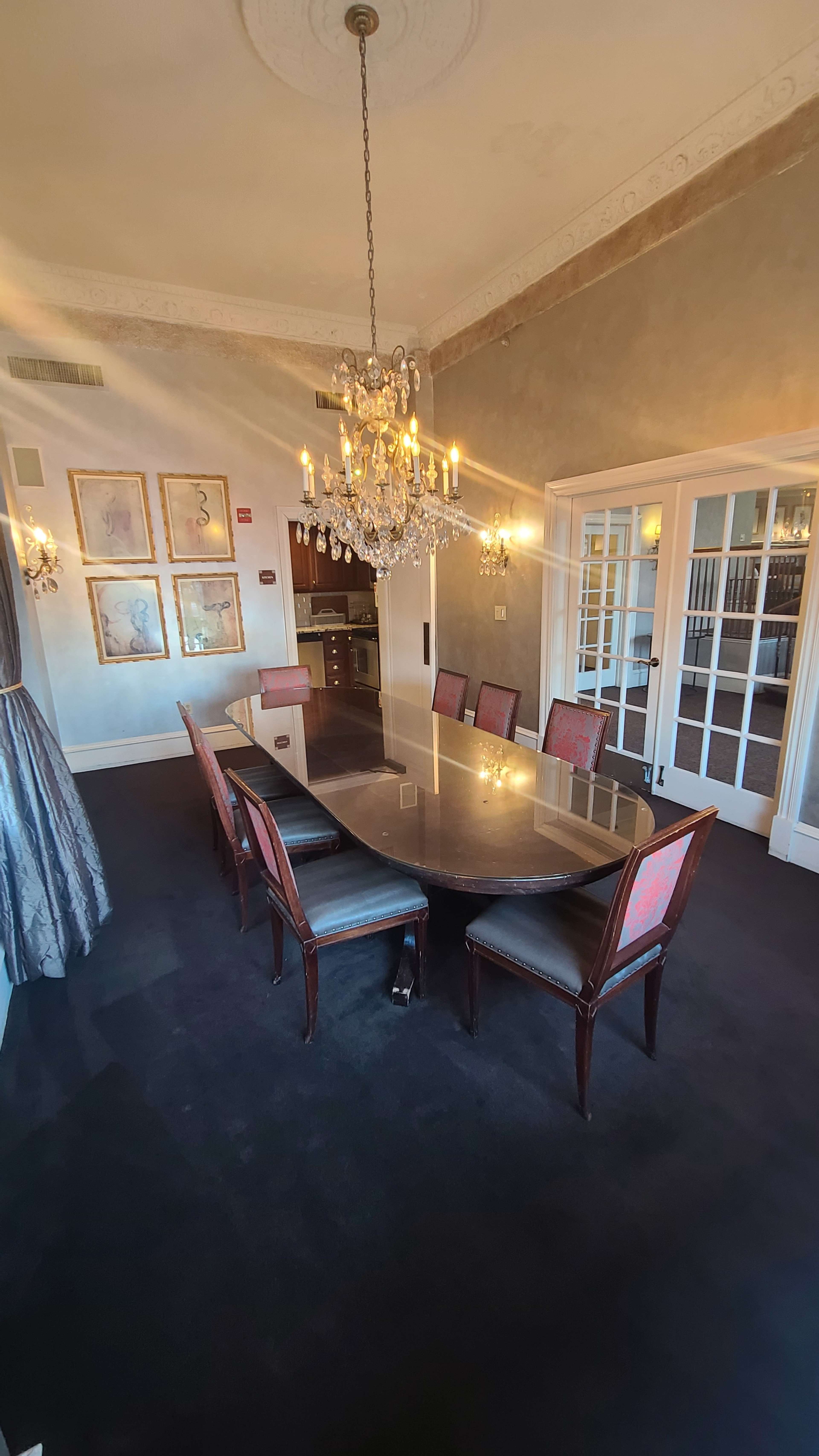 A long, oval table surrounded by seven chairs is set in a well-lit dining room decorated with framed artwork and a chandelier.