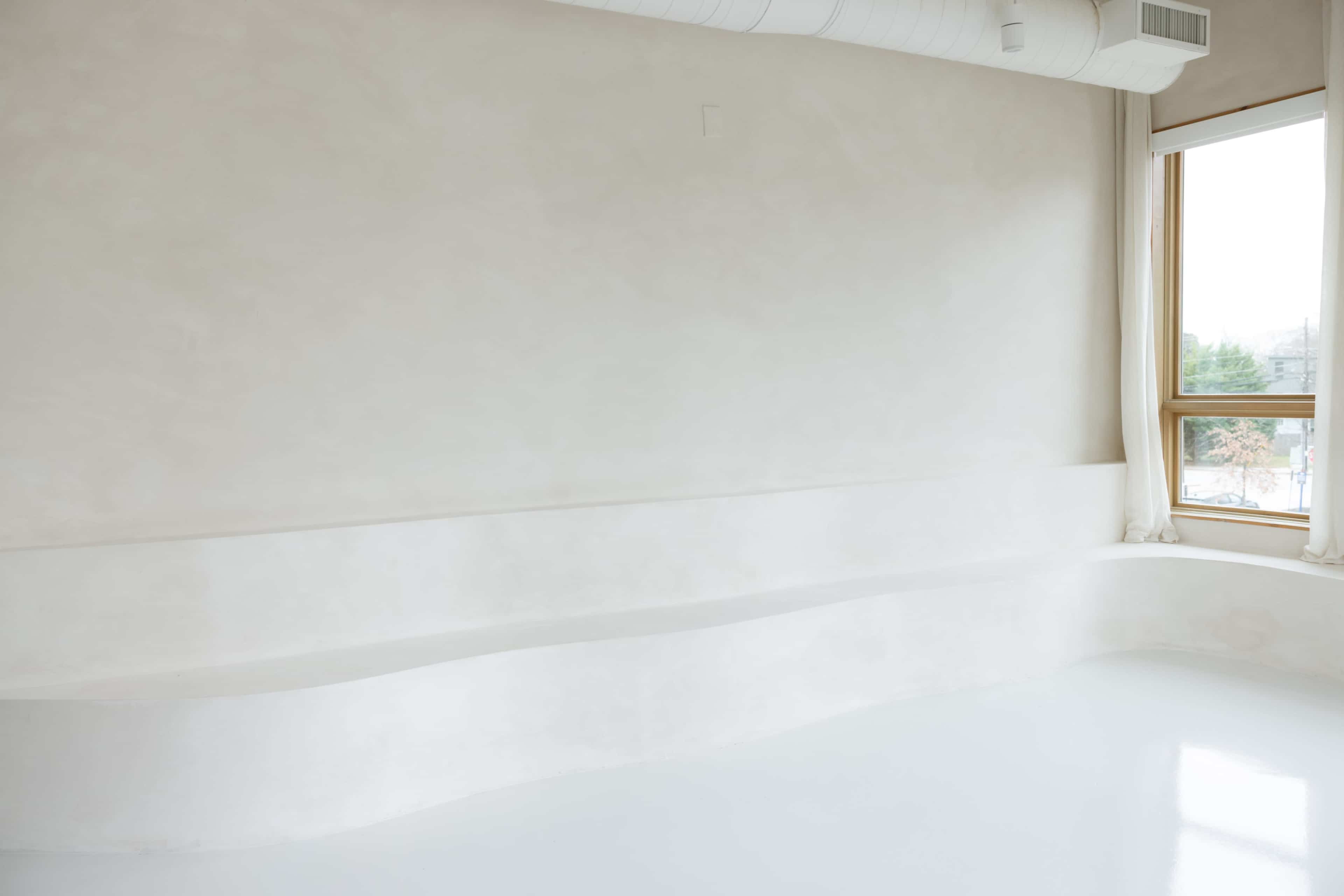 The image shows a minimalist interior with a smooth white wall, a curved white bench, and large windows allowing natural light to enter.