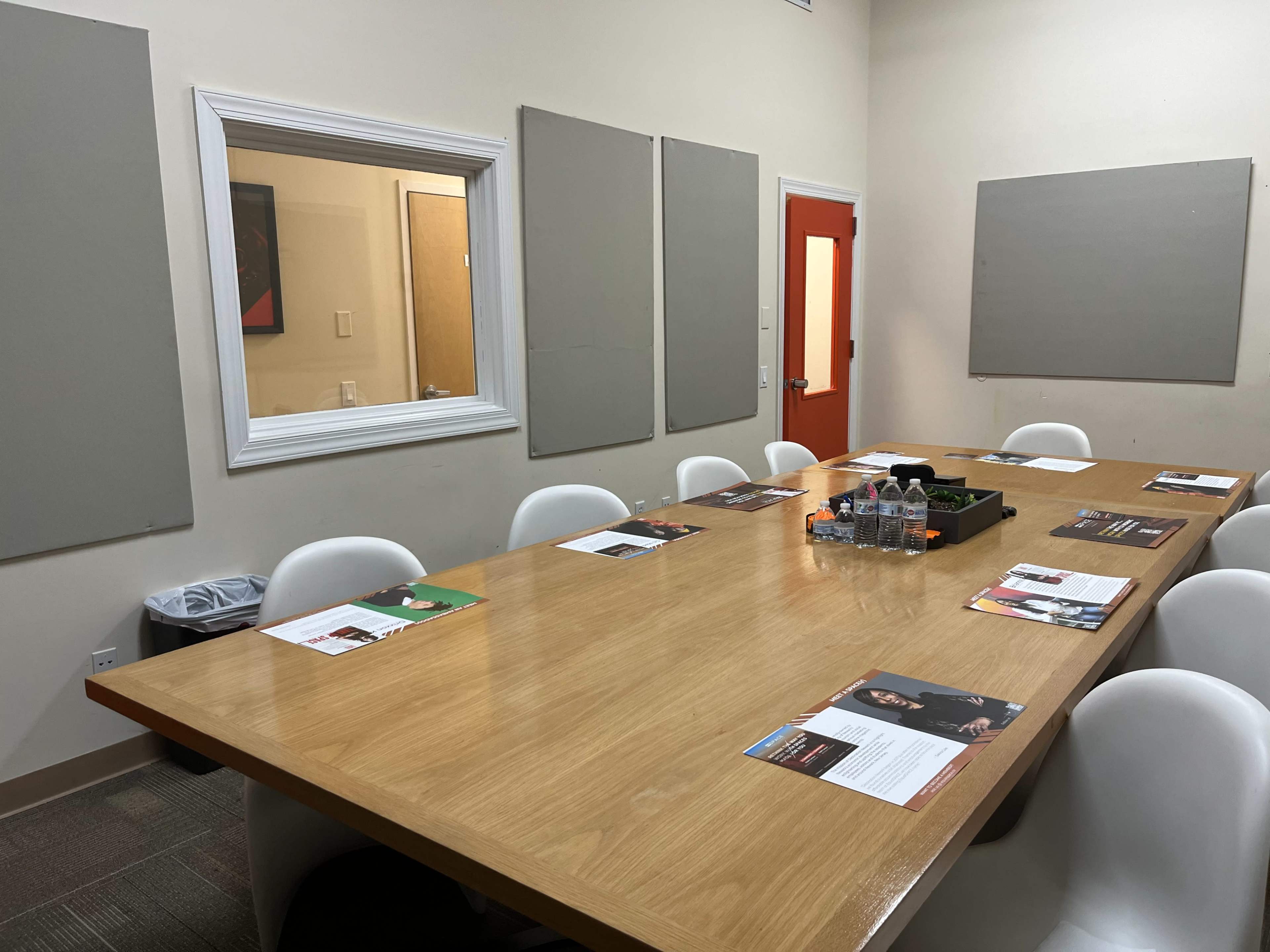 The image shows a conference room with a large wooden table surrounded by white chairs, featuring placemats and water bottles on the table, along with grey soundproof panels on the walls.