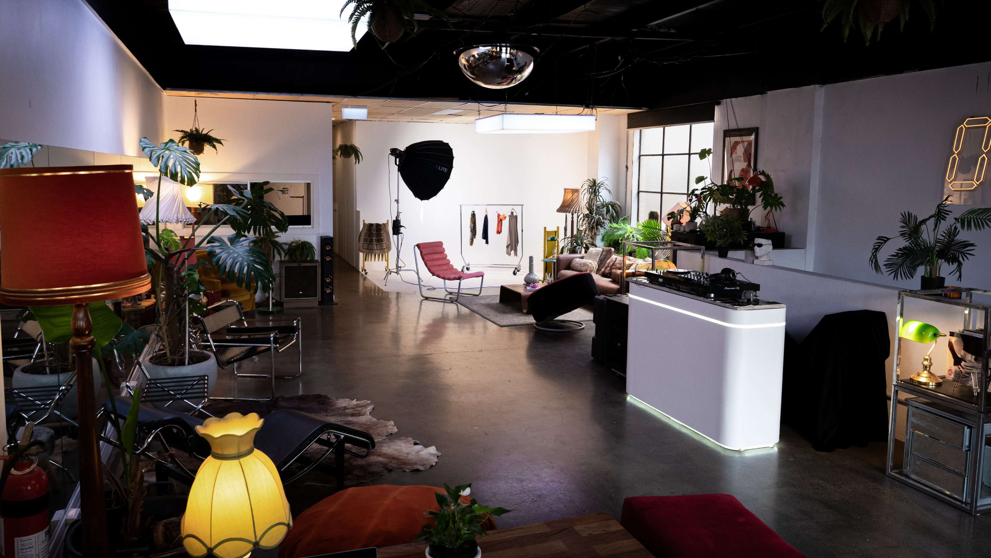 The image shows an interior space designed for a photo shoot, featuring various seating arrangements, plants, lighting equipment, and a bar area.