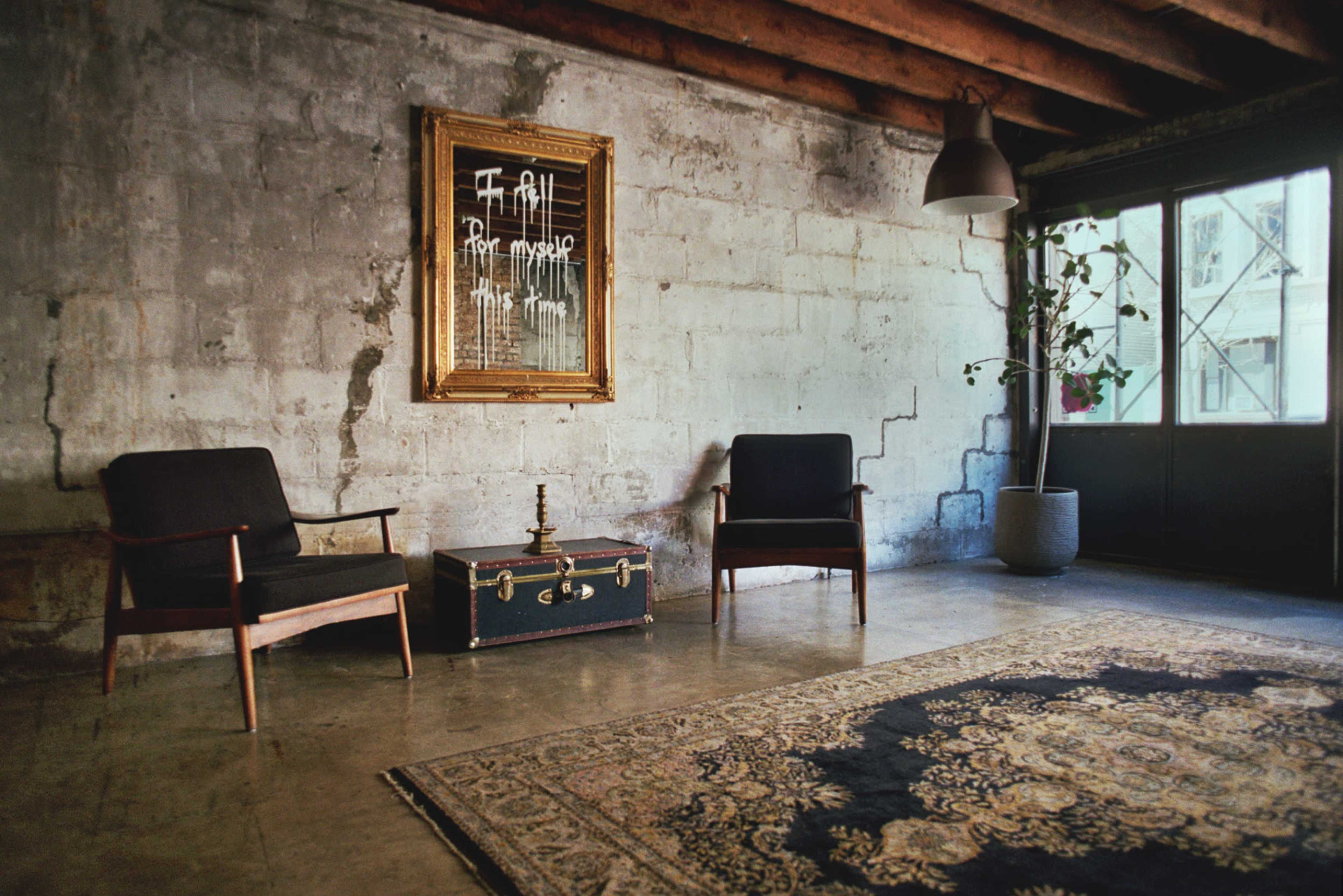 The room features two black chairs facing a vintage trunk, with a framed mirror and a potted plant in a corner, against a concrete wall.
