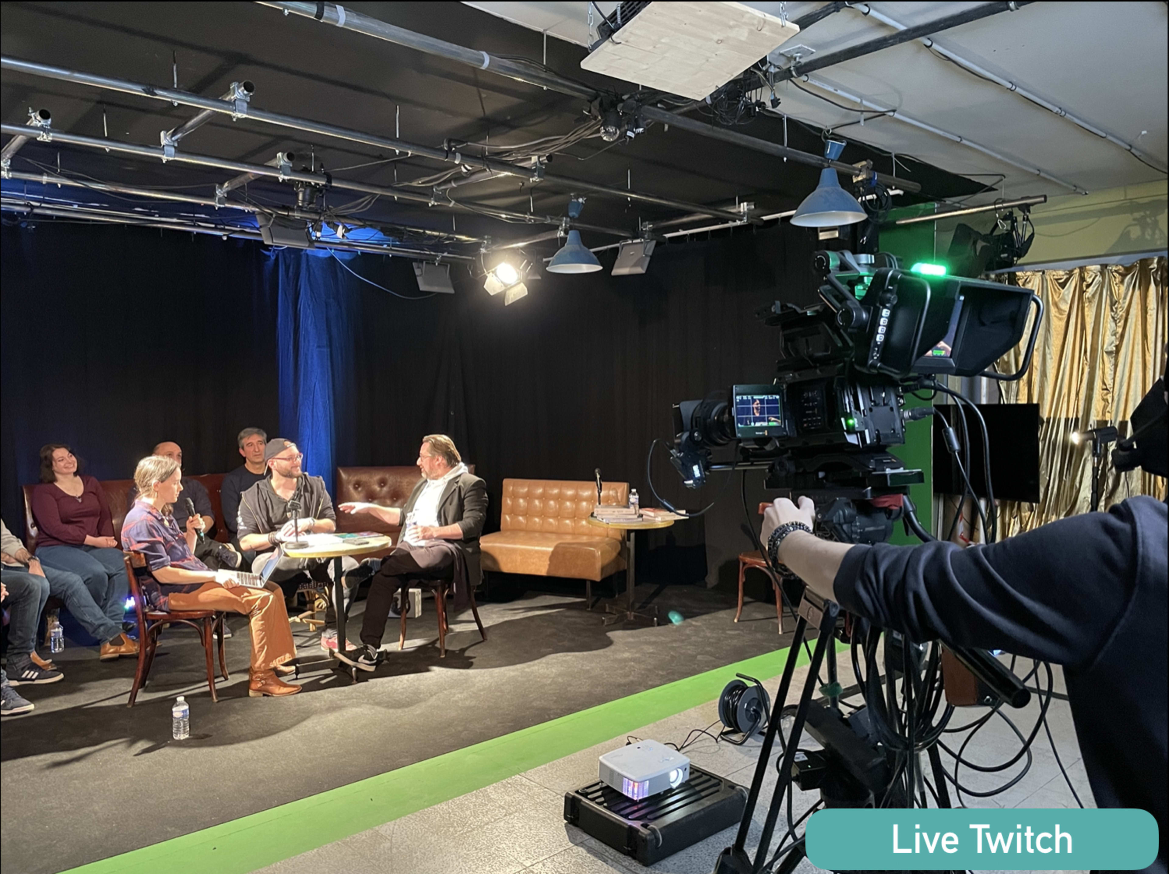 A live Twitch event is taking place on stage with a camera focused on a group of people seated and engaged in conversation.