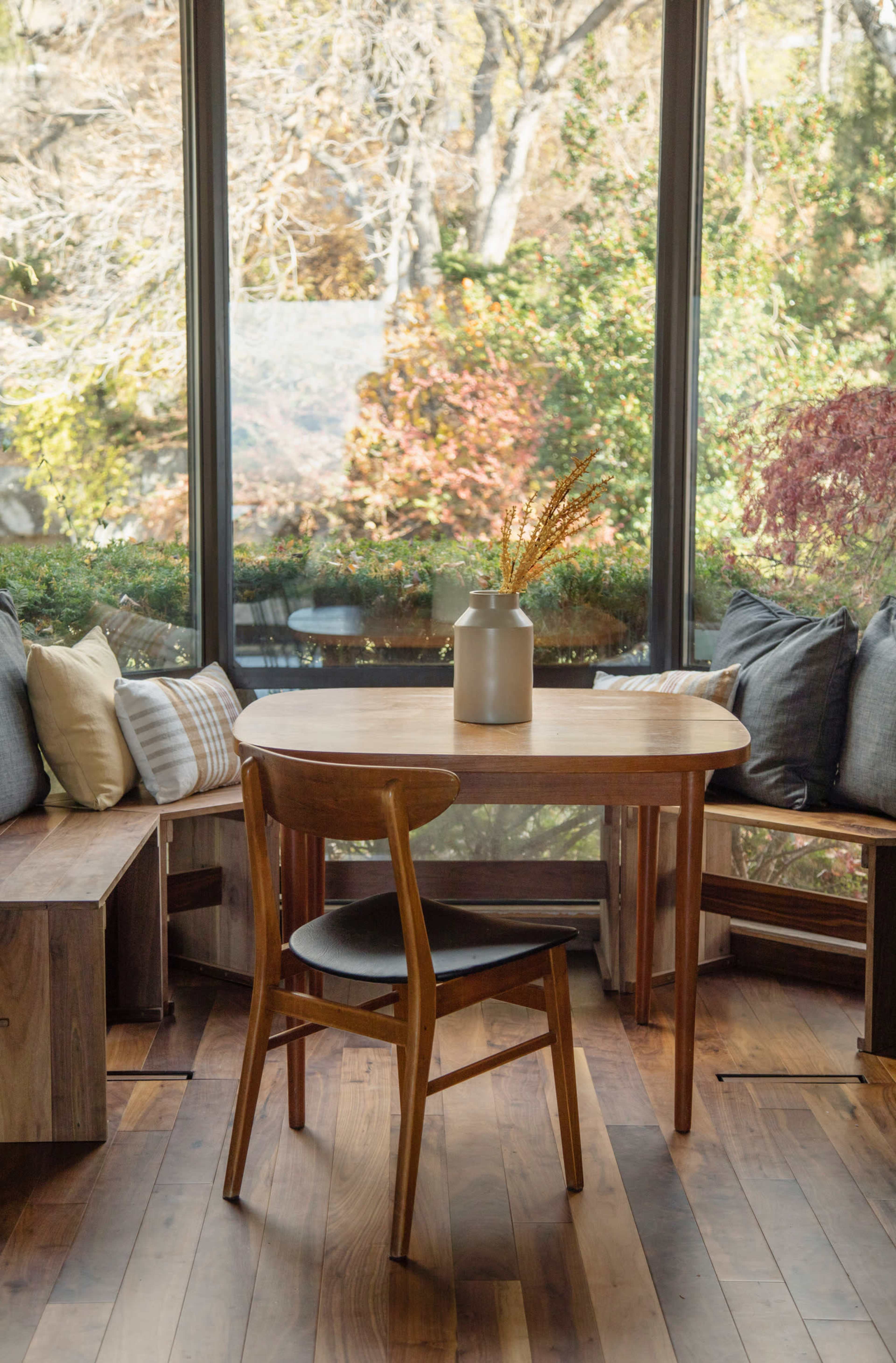 A wooden table is positioned in a sunlit corner of a room with large windows and cozy seating, surrounded by plants and decorative pillows.
