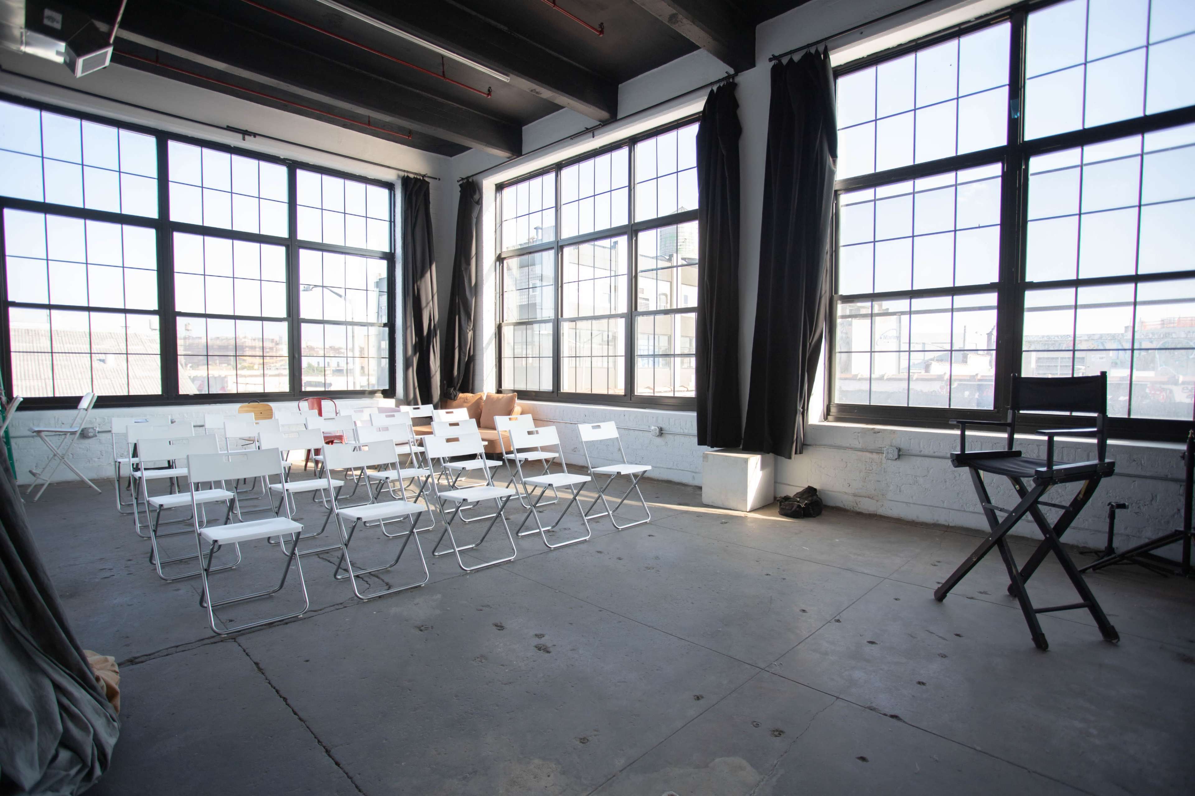 A spacious room with large windows, featuring several white folding chairs arranged in rows and a director's chair positioned in the corner.