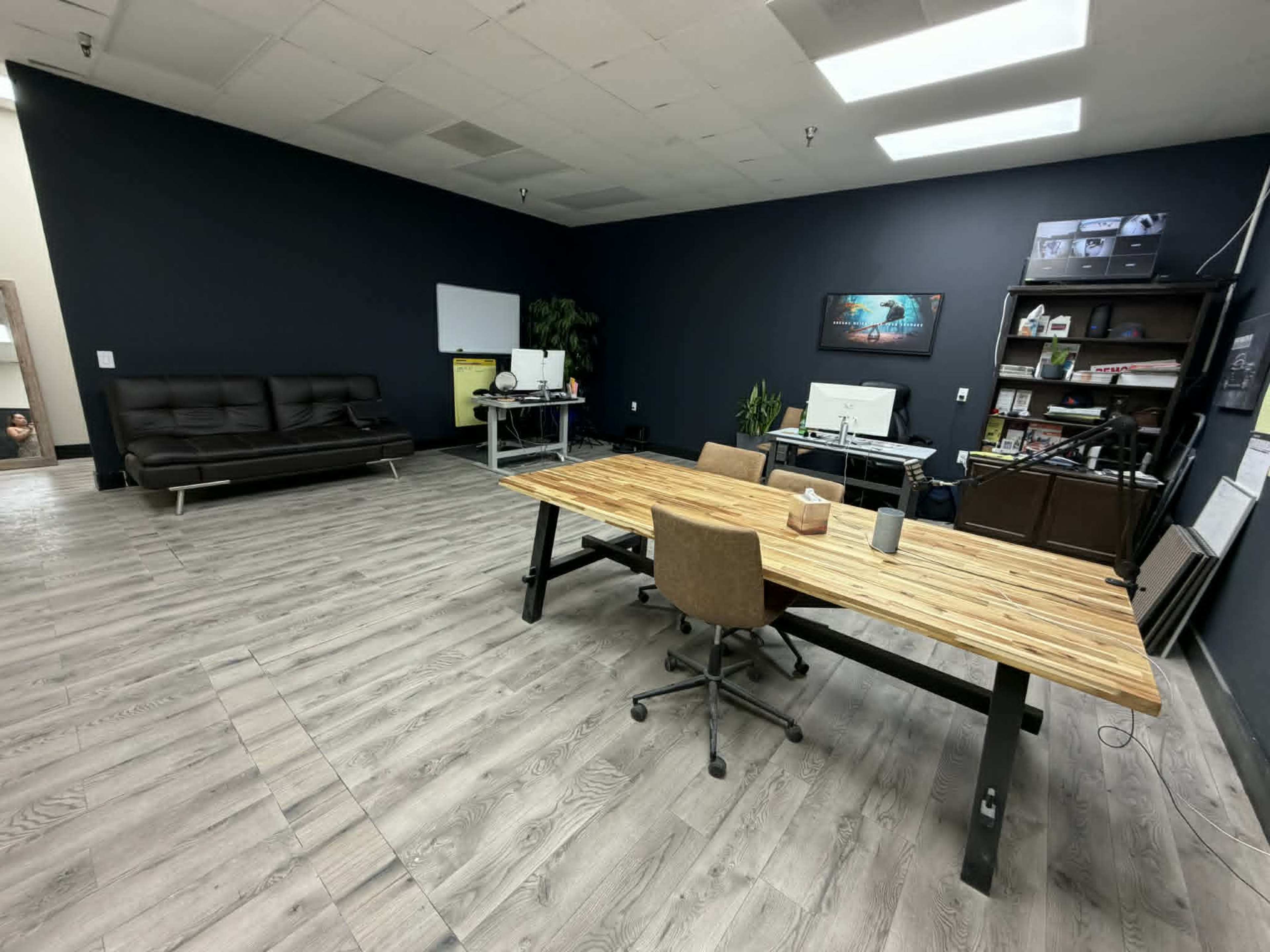 The image shows a spacious office with a wooden table, office chairs, a black couch, a whiteboard, and various workstations along one wall.