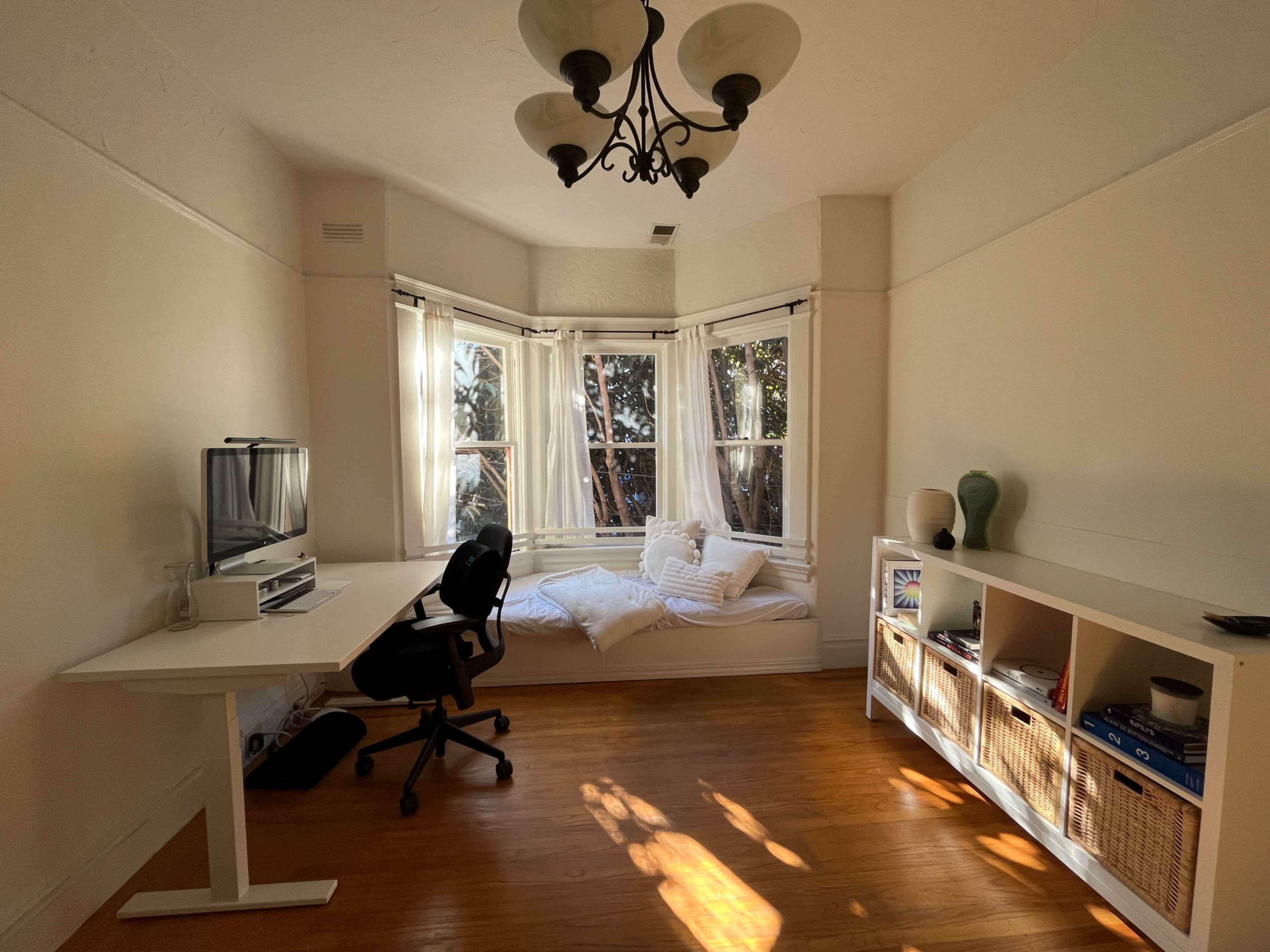 A bright room features a desk with a chair, a wall-mounted television, and a cozy window seat with cushions.