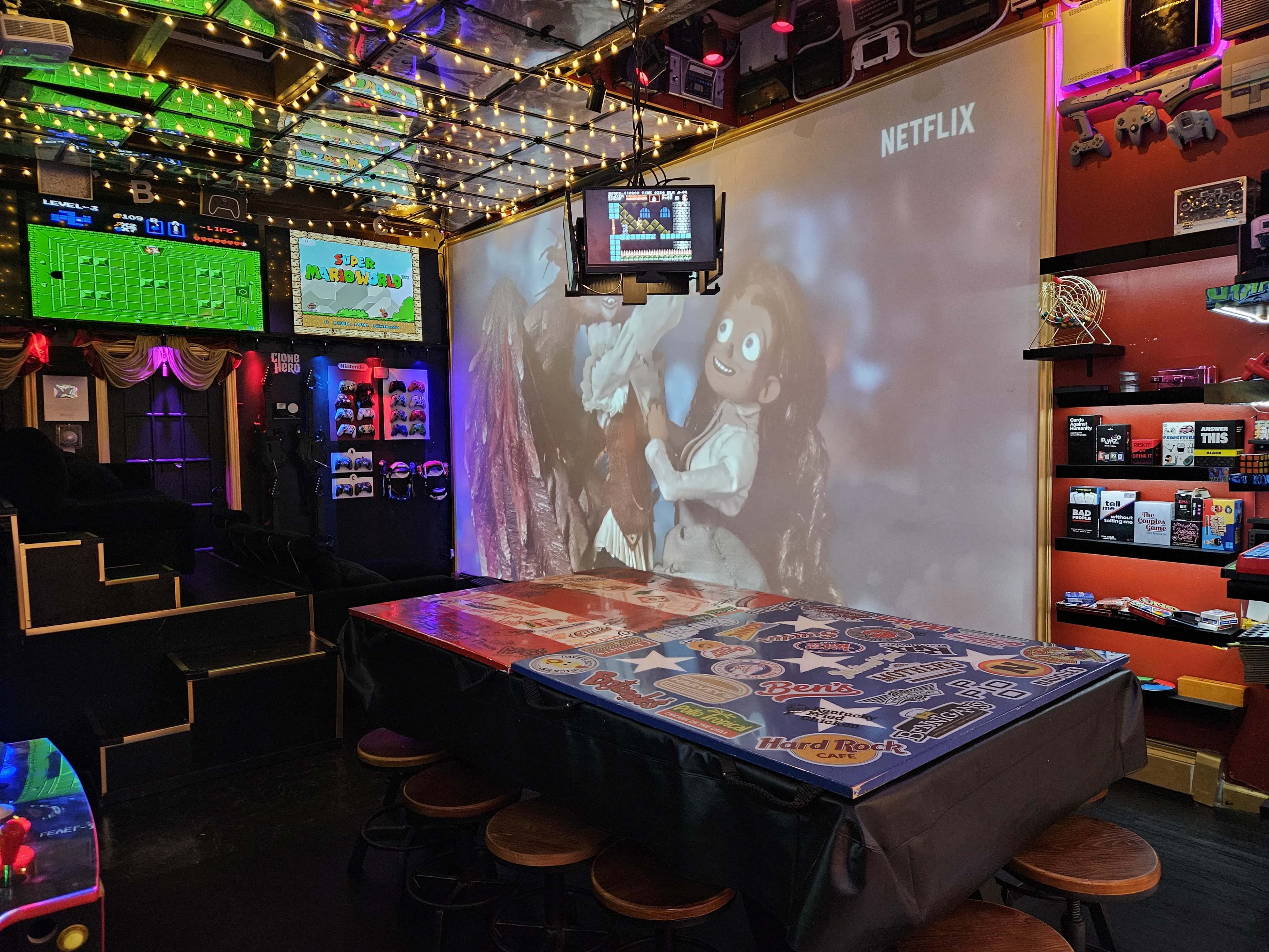 The image shows a lively gaming and entertainment space featuring a table covered in colorful stickers, a screen displaying Netflix content, and retro video game decorations on the walls.