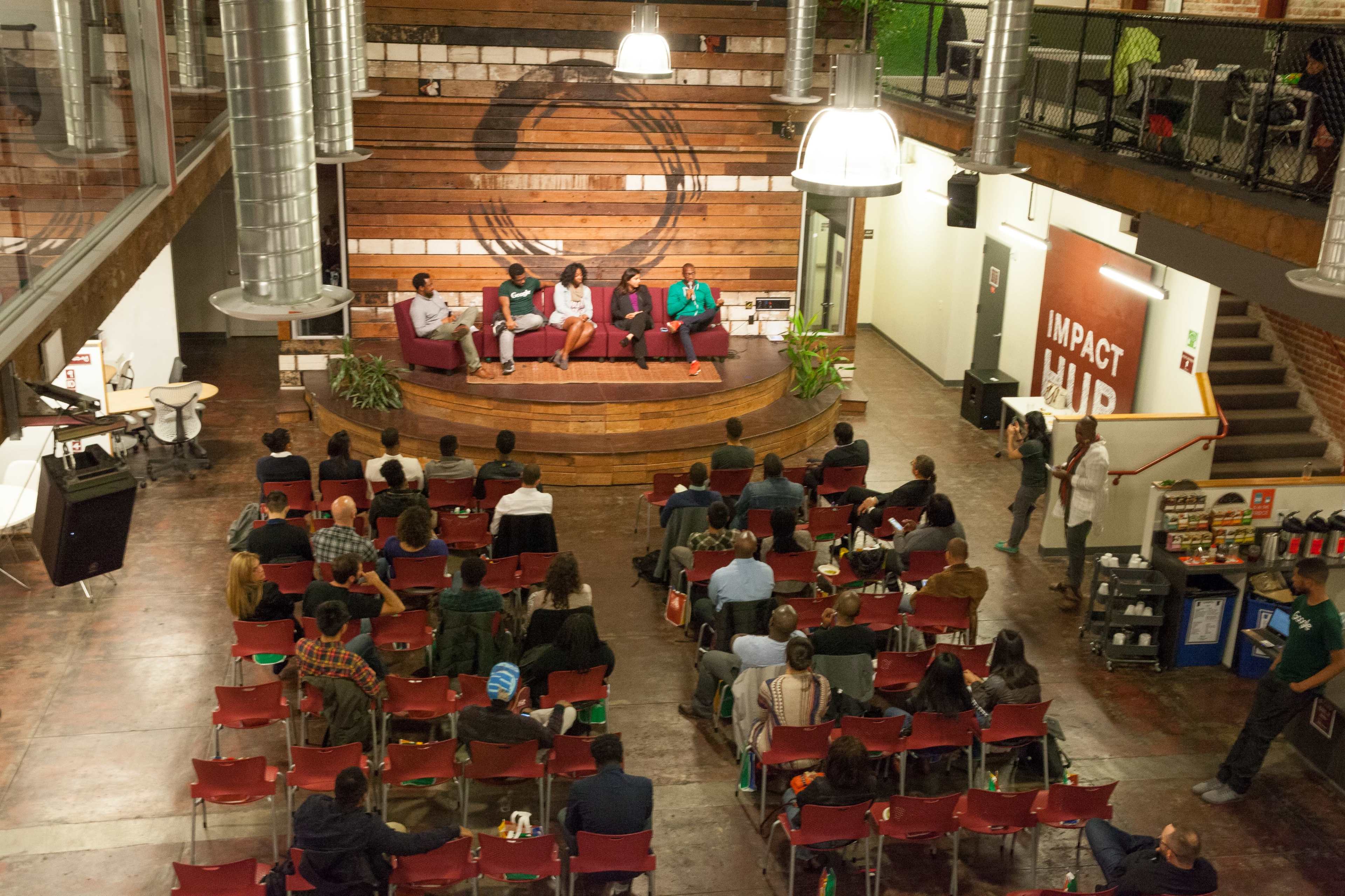 A group of speakers sits on a stage in a large room filled with an audience, surrounded by wooden walls and modern decor.